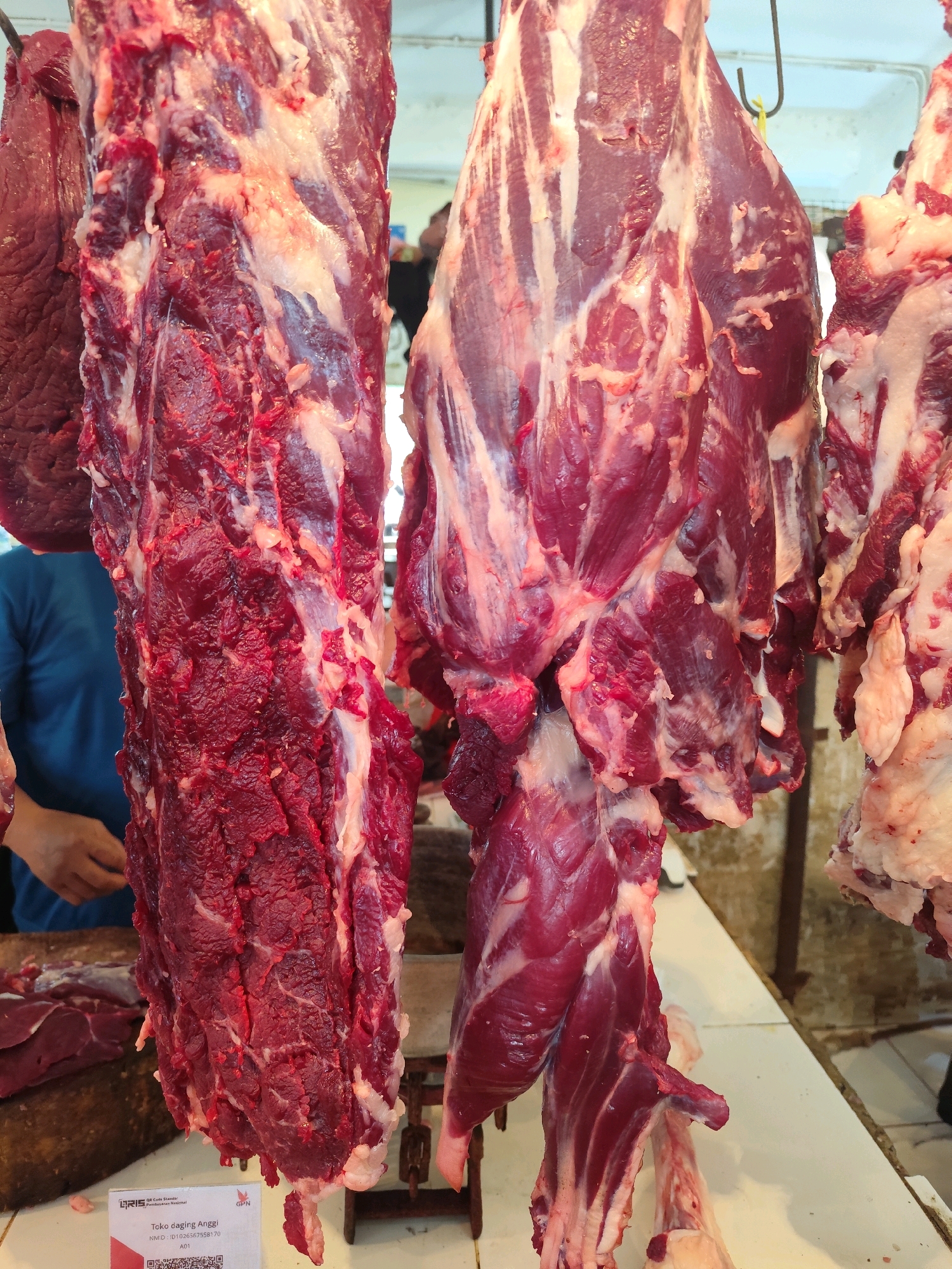 In a dimly lit indoor market, likely a butcher shop in Bogor, Indonesia, several large cuts of raw, red meat hang from hooks. The meat appears to be beef, with visible fat and muscle striations. The cuts are substantial, one particularly large piece dominates the left side of the frame, showing a rough, textured surface. To the right, another large piece hangs, displaying a more layered appearance of muscle and fat. In the background, a person wearing a blue shirt is partially visible, their hands seemingly engaged in preparing or arranging the meat. A small white tag with Indonesian text and a QR code is attached to the lower left of the frame, suggesting a retail environment. The overall impression is one of a bustling marketplace, with the raw meat as the central focus.
