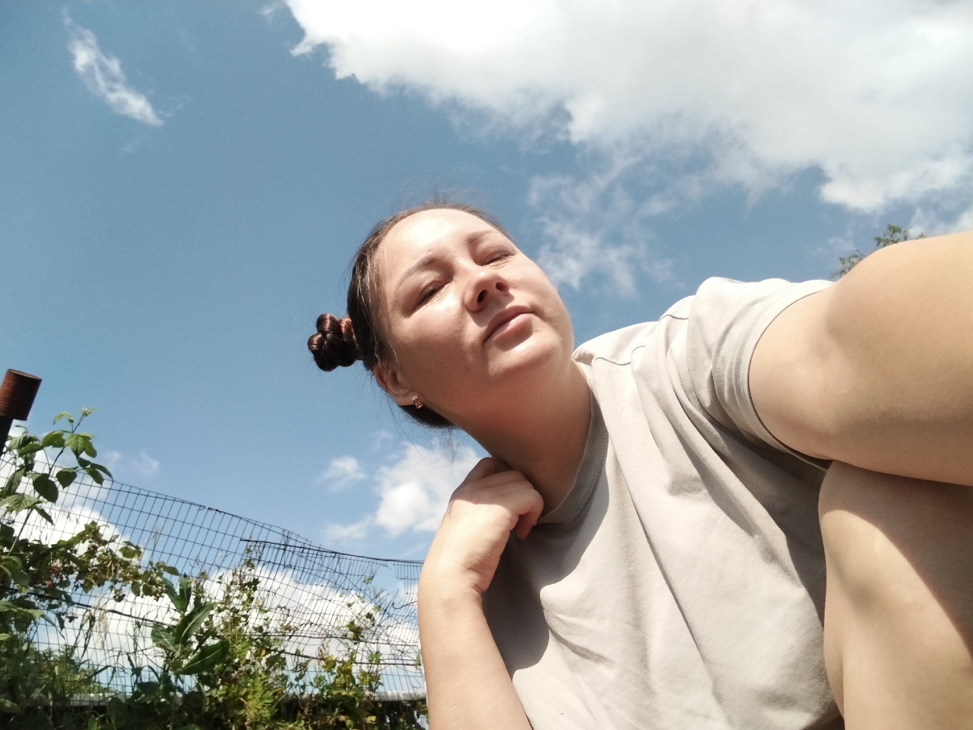 A low-angle, outdoor, mid-shot selfie captures a young woman posing against a bright blue sky with scattered white clouds. She is wearing a light-colored t-shirt and has her dark hair styled in two buns. Her left arm is bent, with her hand resting on her neck, and her eyes are closed, suggesting relaxation. In the background, a chain-link fence and green foliage are visible. The scene is set in a rural area, possibly a backyard or garden. The image appears to have been taken in the village of Vazerki, Russia.