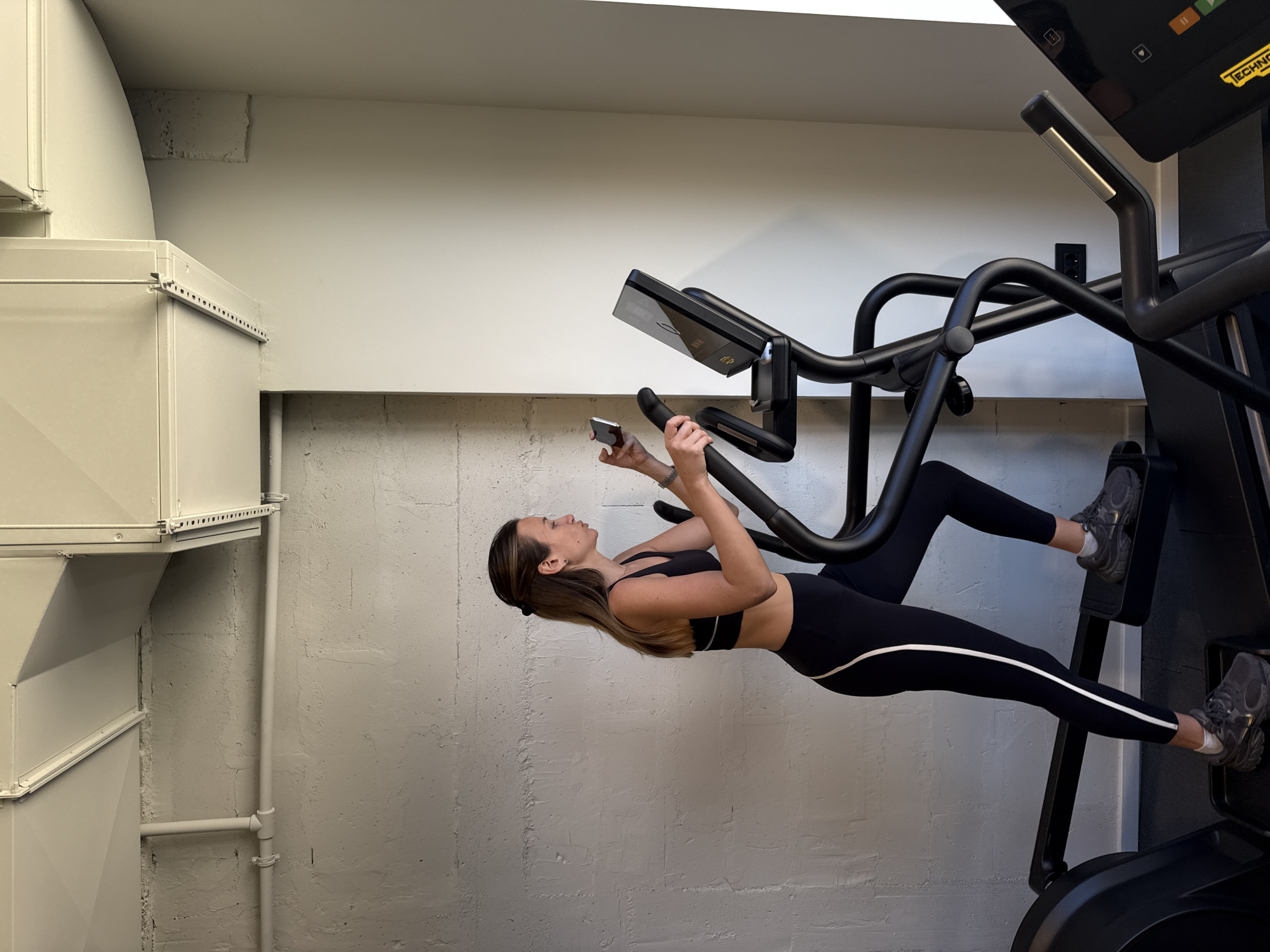 The image shows a woman exercising on an elliptical machine in an indoor setting. The machine is black and features a digital display. The woman is wearing a black sports bra and leggings with a white stripe down the side. She is holding a smartphone and looking at it. To the left of the machine is a large, rectangular metal structure, possibly part of the building's ventilation or ductwork system. The wall behind the machine is textured and painted white. A pipe runs vertically along the wall and connects to a horizontal pipe near the floor. The location is Unknown, Unknown.