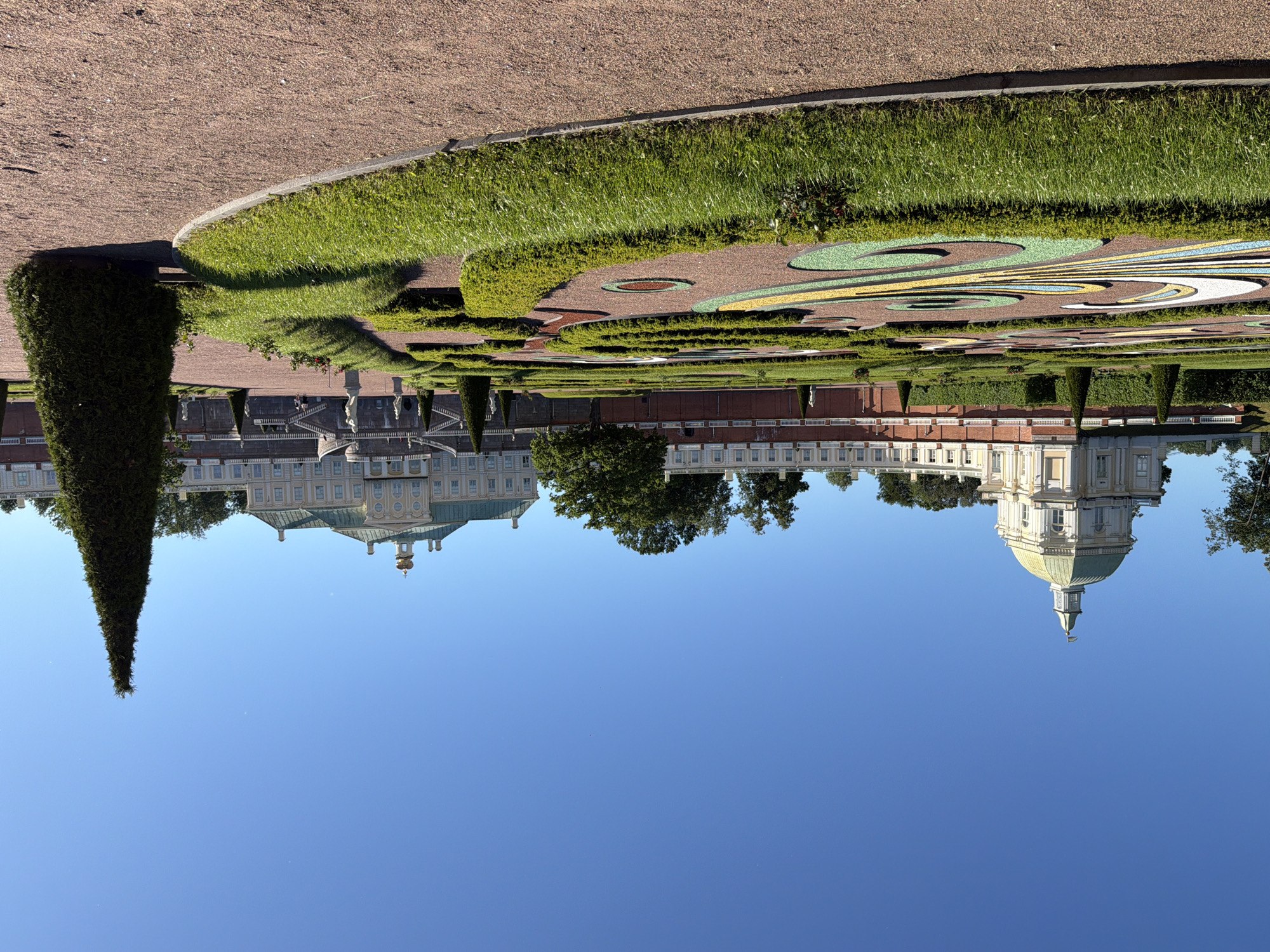 This is a photograph of a garden and a palace complex. The foreground features a meticulously maintained garden with a gravel path, manicured green hedges forming geometric patterns, and a large conical evergreen tree. Beyond the garden, a grand palace stretches across the frame. The palace is multi-storied with a light-colored facade, green roofs, and classical architectural elements. A prominent dome with a golden finial is visible on one end of the palace. Trees are interspersed throughout the grounds. The sky is a clear, bright blue, suggesting a sunny day. The image is oriented vertically, with the sky appearing at the bottom and the ground at the top, indicating it was taken with the camera rotated. No people are visible, and no text is present. The scene evokes a sense of historical grandeur and serene beauty.