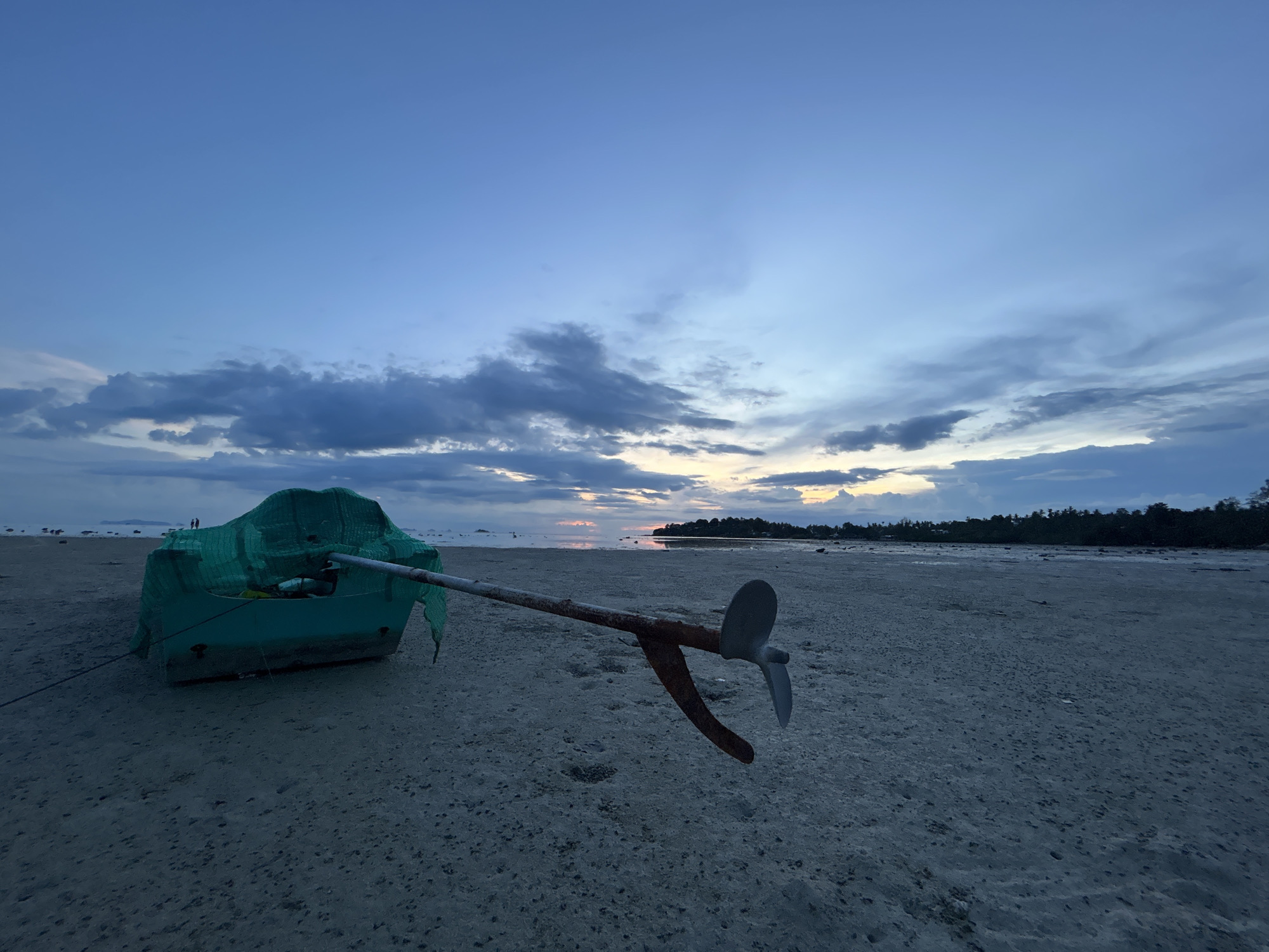 The image depicts a serene beach scene at dusk on Koh Phangan, Thailand. In the foreground, a green boat is partially covered with a green tarp, resting on the sandy shore. An anchor, with its rusty metal shaft and a silver propeller-like flukes, lies extended from the boat, partially obscuring it and pointing towards the right of the frame. The sand is textured with small pebbles and scattered debris.
In the mid-ground, the shallow sea reflects the soft hues of the setting sun. A distant tree line marks the horizon, with silhouettes of palm trees suggesting a tropical environment. A few small islands or sandbars are visible in the distance.
The sky is a canvas of deepening blues and grays, with scattered clouds tinged with the warm colors of sunset near the horizon. The overall mood is peaceful and tranquil, evoking a sense of calm at the end of the day. The time of day is clearly indicated by the twilight sky and the soft, diffused light. There is no visible text in the image.