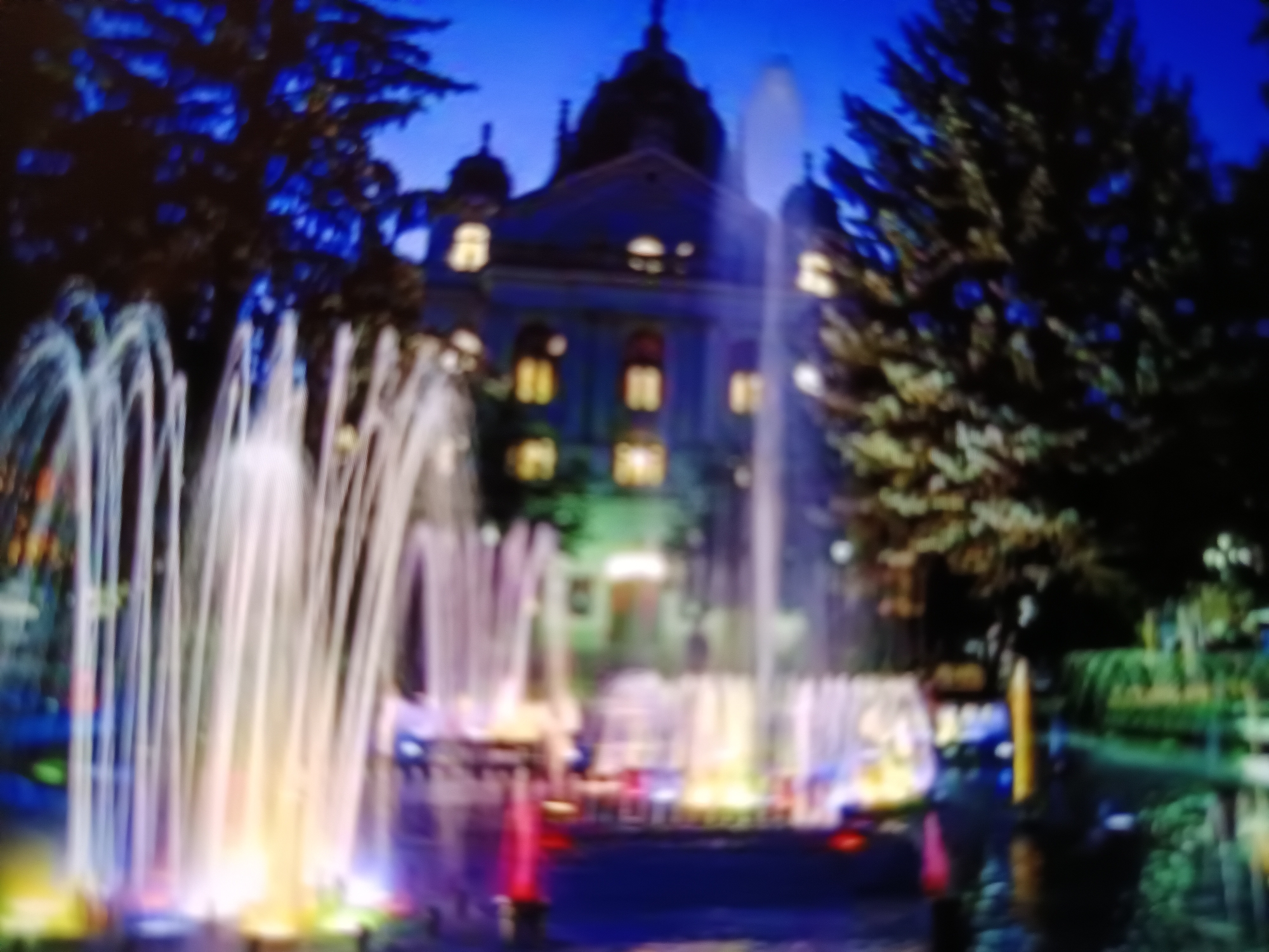 The image shows a fountain illuminated with colorful lights at night in front of a building. The building appears to be a historical structure with domes and multiple windows emitting a warm yellow light. Trees flank the building, and the sky is a deep blue, suggesting twilight or nighttime. The water jets of the fountain are brightly lit, creating a vibrant and dynamic display. The scene is likely a public square or park, and the architectural style of the building and the fountain suggest it could be a prominent landmark. Specifically, this image depicts the Singing Fountain in front of the State Theatre in Košice, Slovakia.