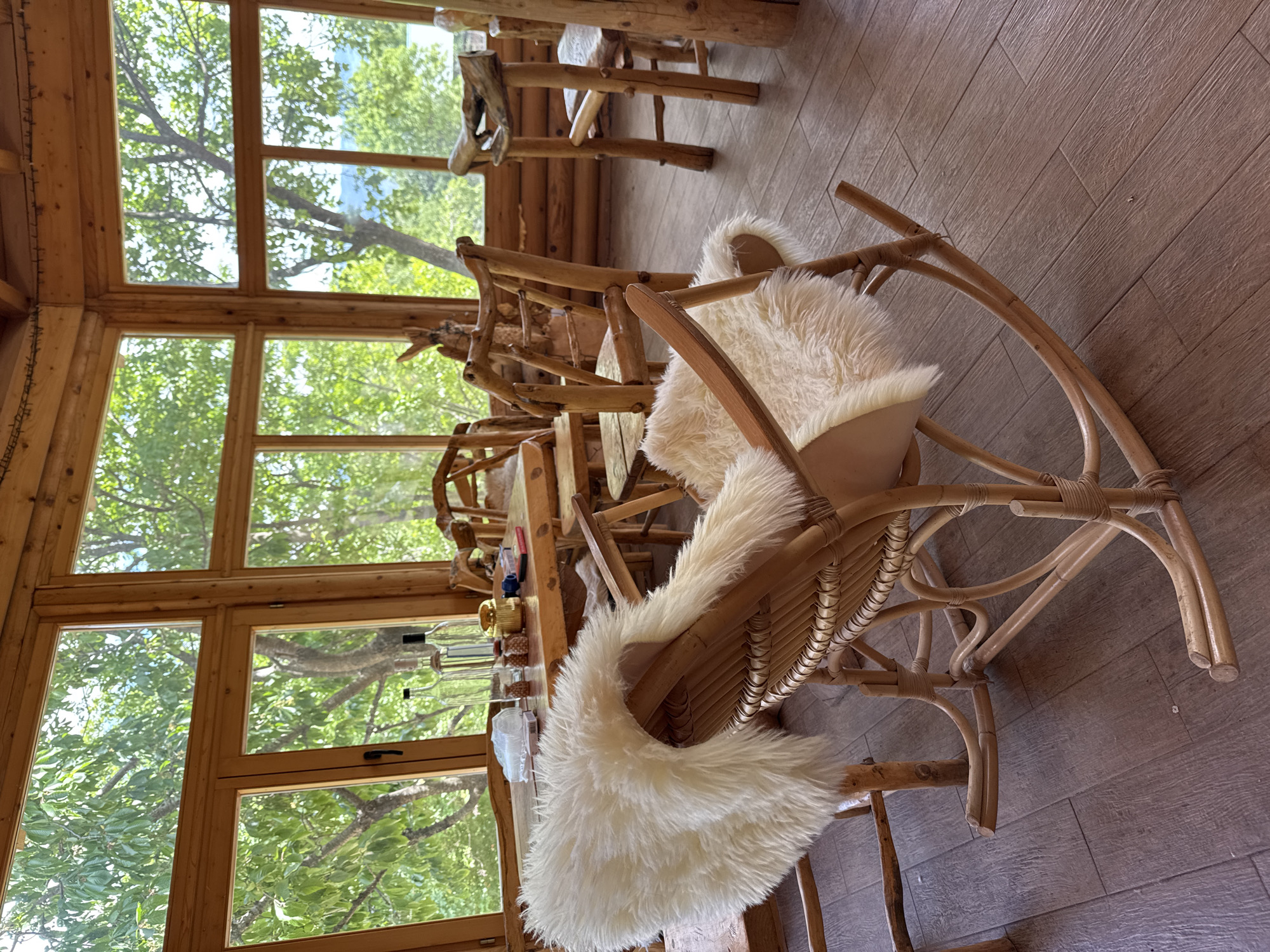 A room in Gornje Mrke, Montenegro, features a wooden table set with glassware and other items. The scene is bright, with natural light streaming in through the window. Several wooden chairs, some with white fur cushions, are arranged around the table. The room appears to be part of a wooden structure, with wooden floors and walls. The view from the window includes trees and foliage.