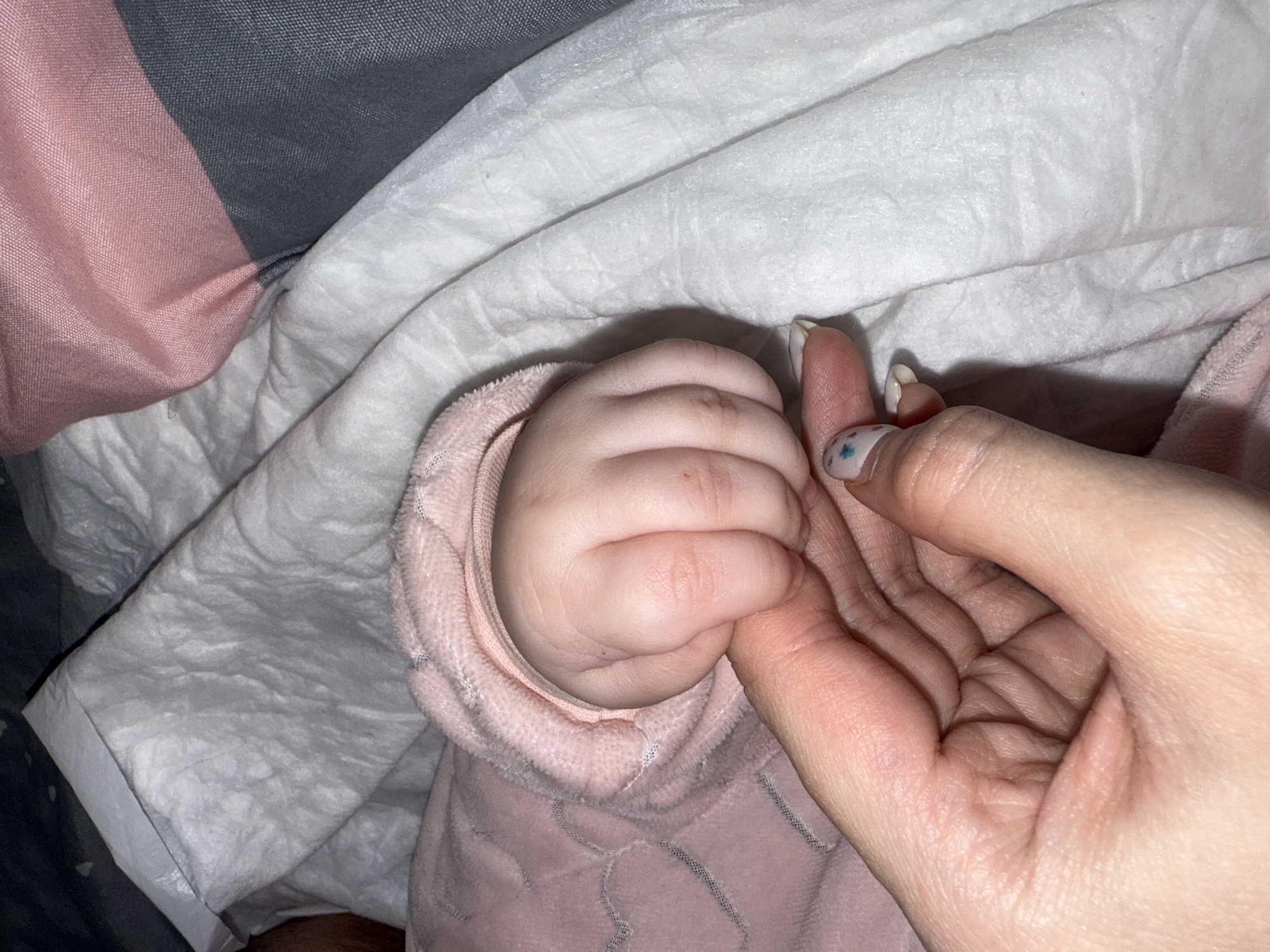 The image captures a tender moment between a baby and an adult, set in Chaikovsky, Russia. The primary subjects are the plump, small hand of an infant tightly grasping the index finger of an adult's hand. The baby's hand is visible from the wrist up, wearing a soft, textured pink garment, possibly a hooded sleeper or blanket, with an embossed or patterned design. The adult's hand, likely a right hand, shows smooth skin and neatly manicured nails; the thumbnail has a light polish with small, colorful star or dot accents.
The scene takes place indoors, likely in a comfortable home setting, with various soft fabrics providing the background. These include a crumpled white sheet or blanket on which the hands rest, a soft grey and pink striped fabric visible in the upper left, and more of the textured pink fabric belonging to the baby's attire. The lighting is soft and ambient, suggesting an intimate moment, possibly during the day or evening. No specific time of day or weather conditions are discernible. The image evokes feelings of warmth, protection, and deep connection between the two individuals. There is no visible text in the image.