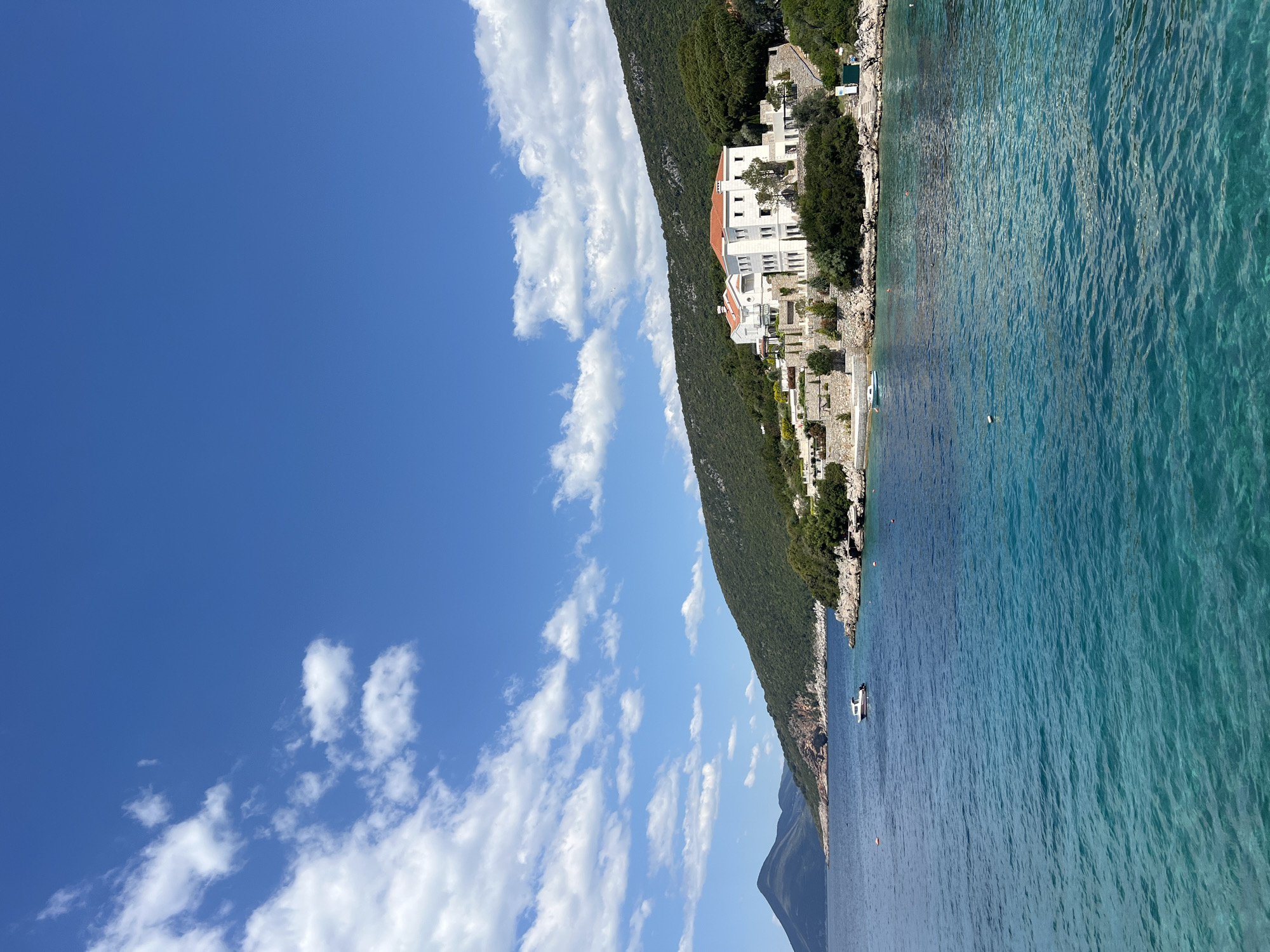 The image shows a shoreline in Luštica, Montenegro, where a building complex sits atop a steep, green slope that meets the sea. A blue sky with scattered clouds covers the upper left portion of the image. The water is clear and blue. A boat is seen on the water.