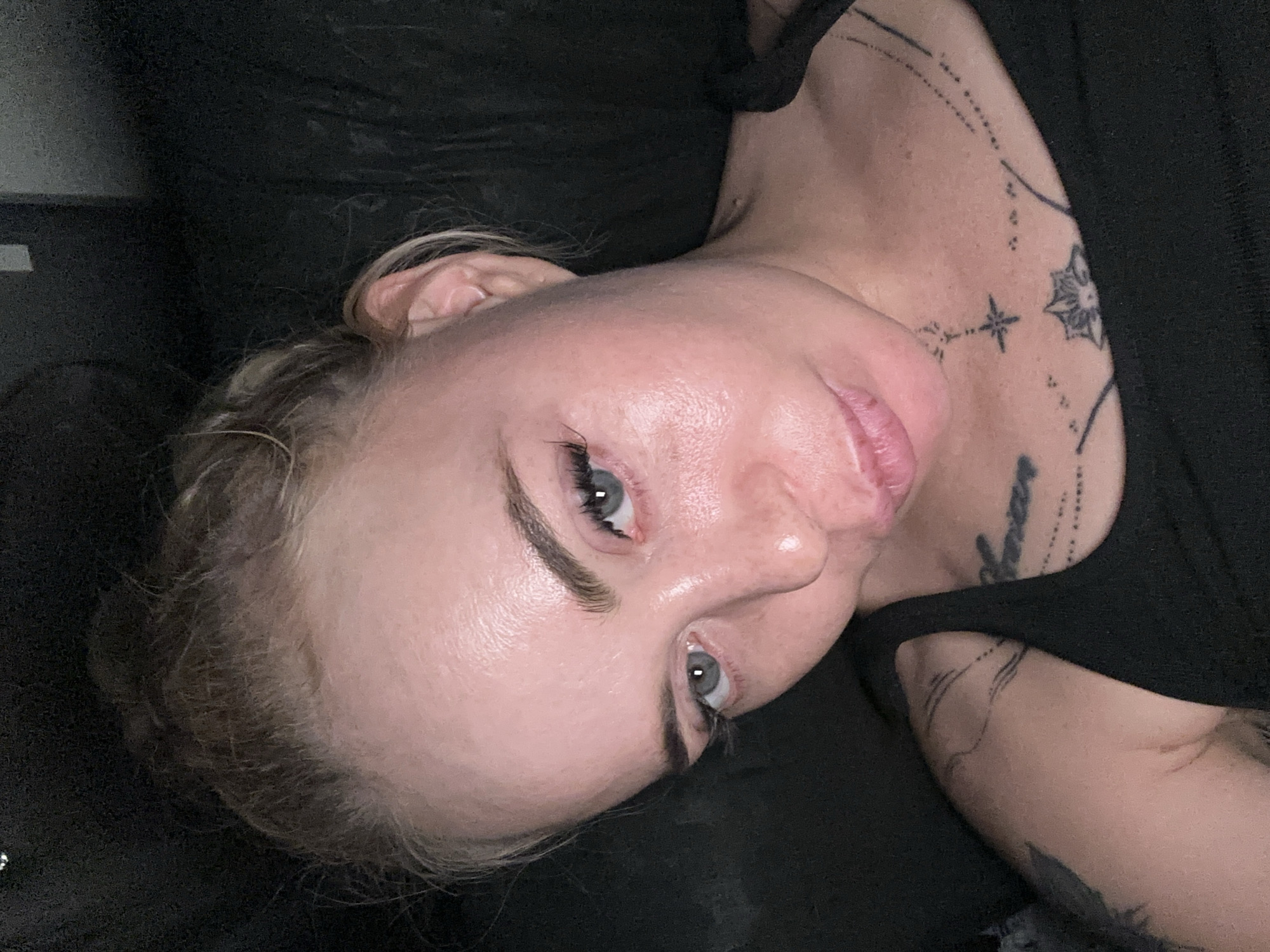 A medium-shot, eye-level, indoor, selfie-style photograph shows a Caucasian woman with blonde hair pulled up into a bun. She is wearing a black tank top with tattoos visible on her chest and left arm. The woman has blue-grey eyes, fair skin with some visible pores and shine, and her lips are slightly parted. The background is dark and indistinct.