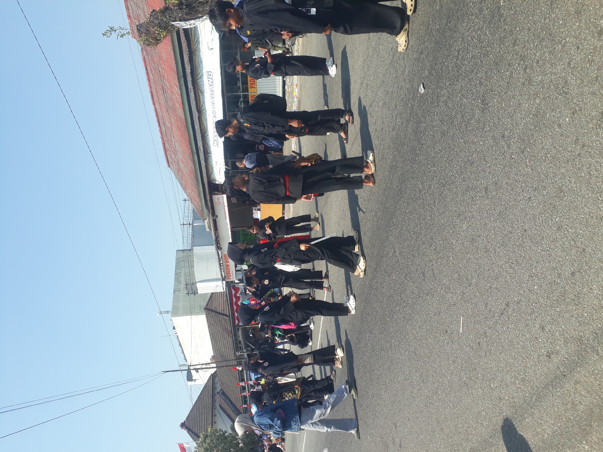 A group of young people, likely students of a martial art, are gathered on a paved street under a clear blue sky. Many are wearing matching black uniforms with red belts. Some are talking, while others appear to be waiting. The background shows buildings with red roofs, one of which has a sign that reads "MASAKAN PADANG" (Padang Cuisine). A small Indonesian flag is visible on a pole to the left. The scene suggests a public event or demonstration in a city setting.