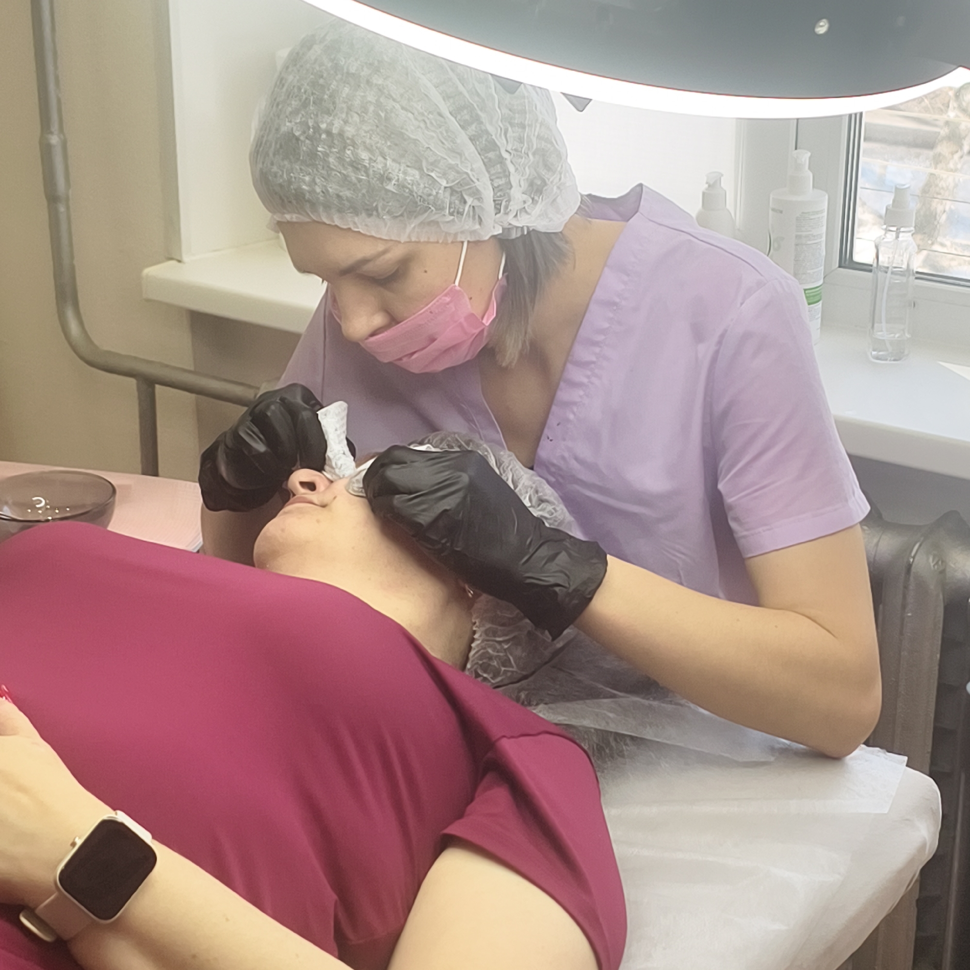 The scene depicts an indoor treatment room, likely a beauty salon or clinic, in the city of Unknown, Unknown. A technician, identifiable by a light purple short-sleeved uniform, white disposable hairnet, pink disposable face mask, and black disposable gloves, is leaning over a client. The technician is performing a procedure on the client's face, specifically near the nose and eye area, using small white disposable pads or wipes held in her gloved hands.
The client is lying supine on a white examination table or bed, which is covered with a white disposable sheet. The client wears a dark magenta-colored top, and a light-colored smartwatch is visible on her left wrist, with red nail polish on her fingertips.
To the left of the client's head on the table, a small, dark glass bowl containing a clear substance is present. In the background, a white window sill holds several items: two white pump bottles of varying sizes and a clear spray bottle. Below the window sill, a dark, cast iron radiator is partially visible. Overhead, a large, rectangular light fixture, likely for illuminating the treatment area, is partially visible. A grey metal pole, possibly part of the light fixture or other equipment, is seen on the left side of the frame. The room walls are a light, neutral color.