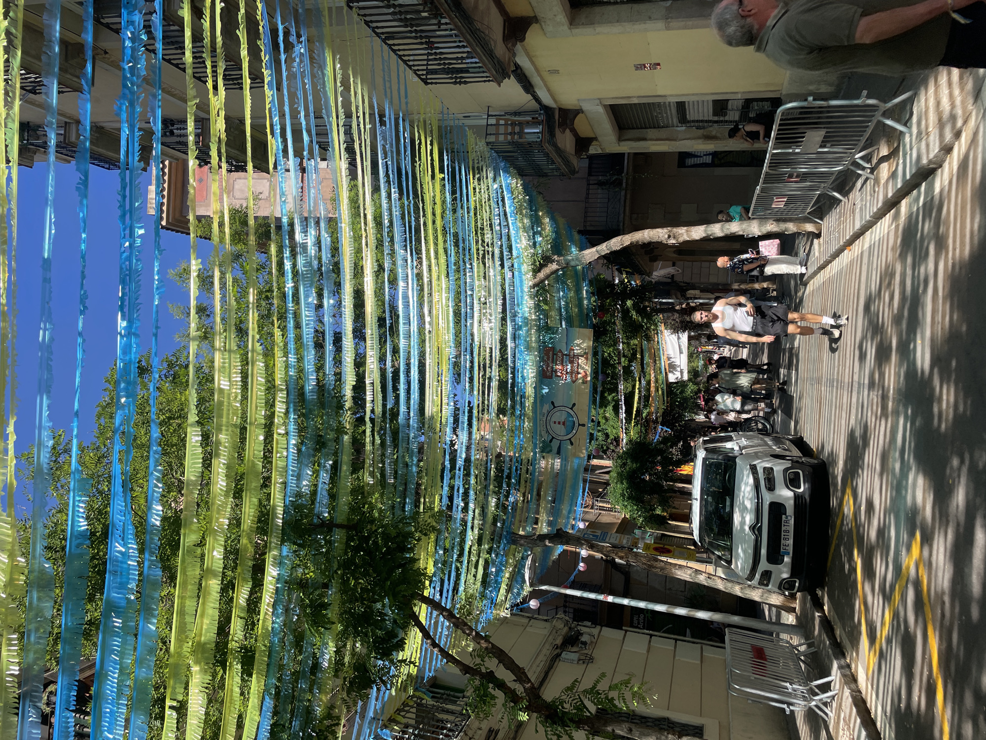 The image captures a street scene, likely in Barcelona, Spain. The street is adorned with vertical hanging decorations of blue and yellow. Pedestrians are visible on the right side of the frame. There is a white car parked on the right side of the street. In the background, some buildings are visible under a clear blue sky. The scene looks like a sunny day.