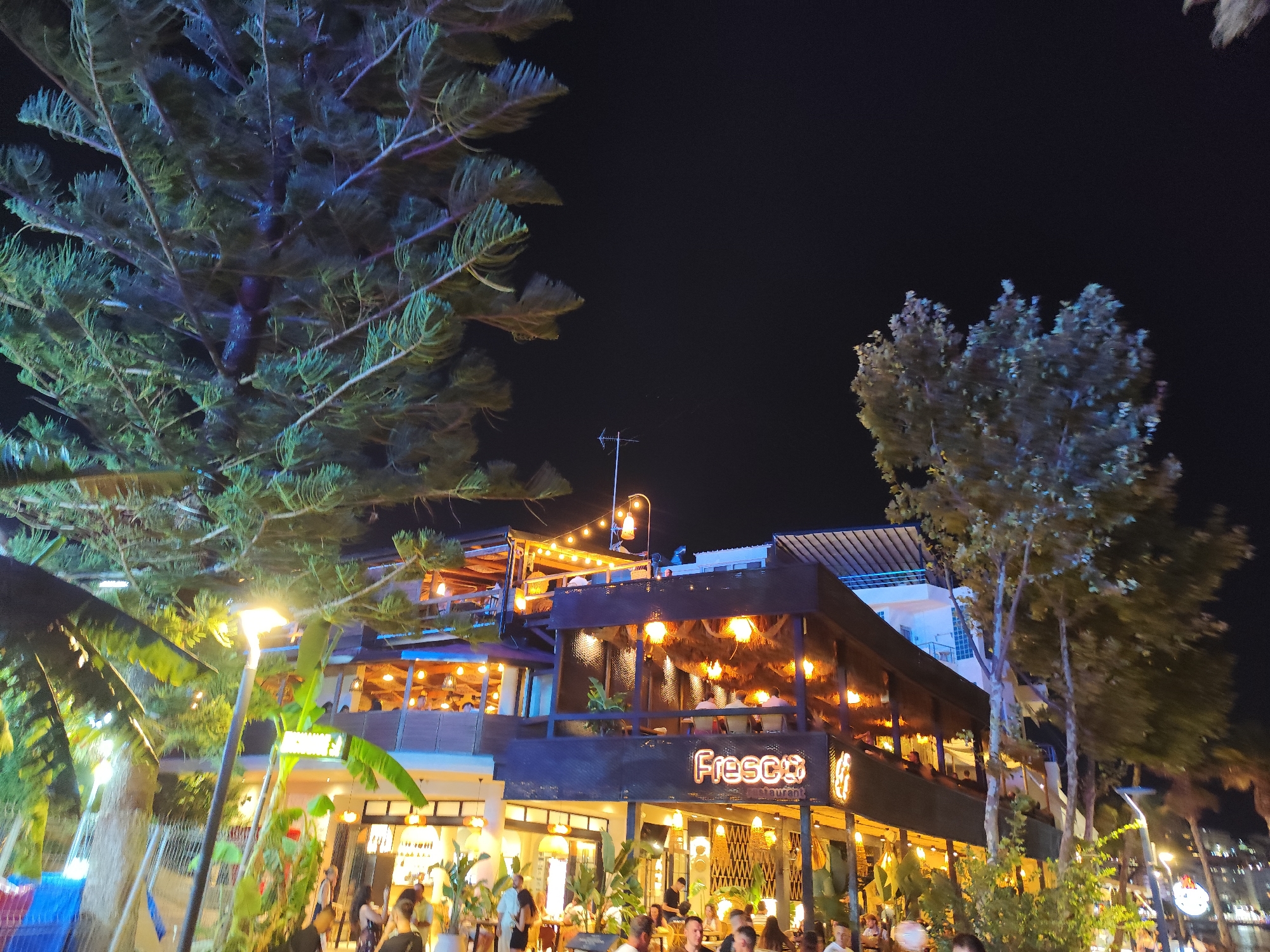 The image depicts a vibrant nighttime scene at a restaurant in Saranda, Albania. The restaurant, named "Fresco," features a multi-level structure with glowing neon signage and numerous ambient lights illuminating the facade and interior. People are visible dining and socializing on the upper and lower levels, as well as on the sidewalk area in front of the establishment. The scene is characterized by lush vegetation, including palm trees and other tropical-looking plants, which are interspersed with streetlights and the restaurant's lighting. The overall atmosphere suggests a lively evening at a popular dining spot.