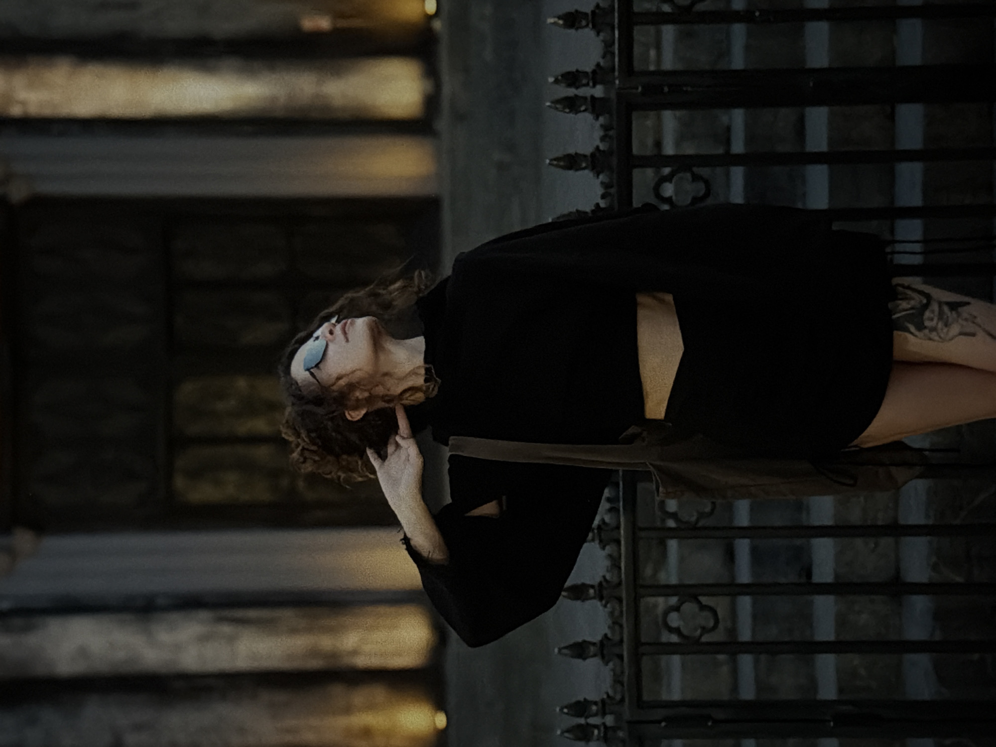 A portrait of a young woman with curly brown hair and glasses, wearing a black dress. She is standing outdoors, possibly in a urban environment, next to an ornate iron fence. Her right hand is raised, and her finger is touching her face near her ear. On her left thigh is visible a tattoo. The setting appears to be a building facade, as suggested by the light reflecting off the brickwork. There is no indication that this photo was taken in Da Nang, Vietnam.