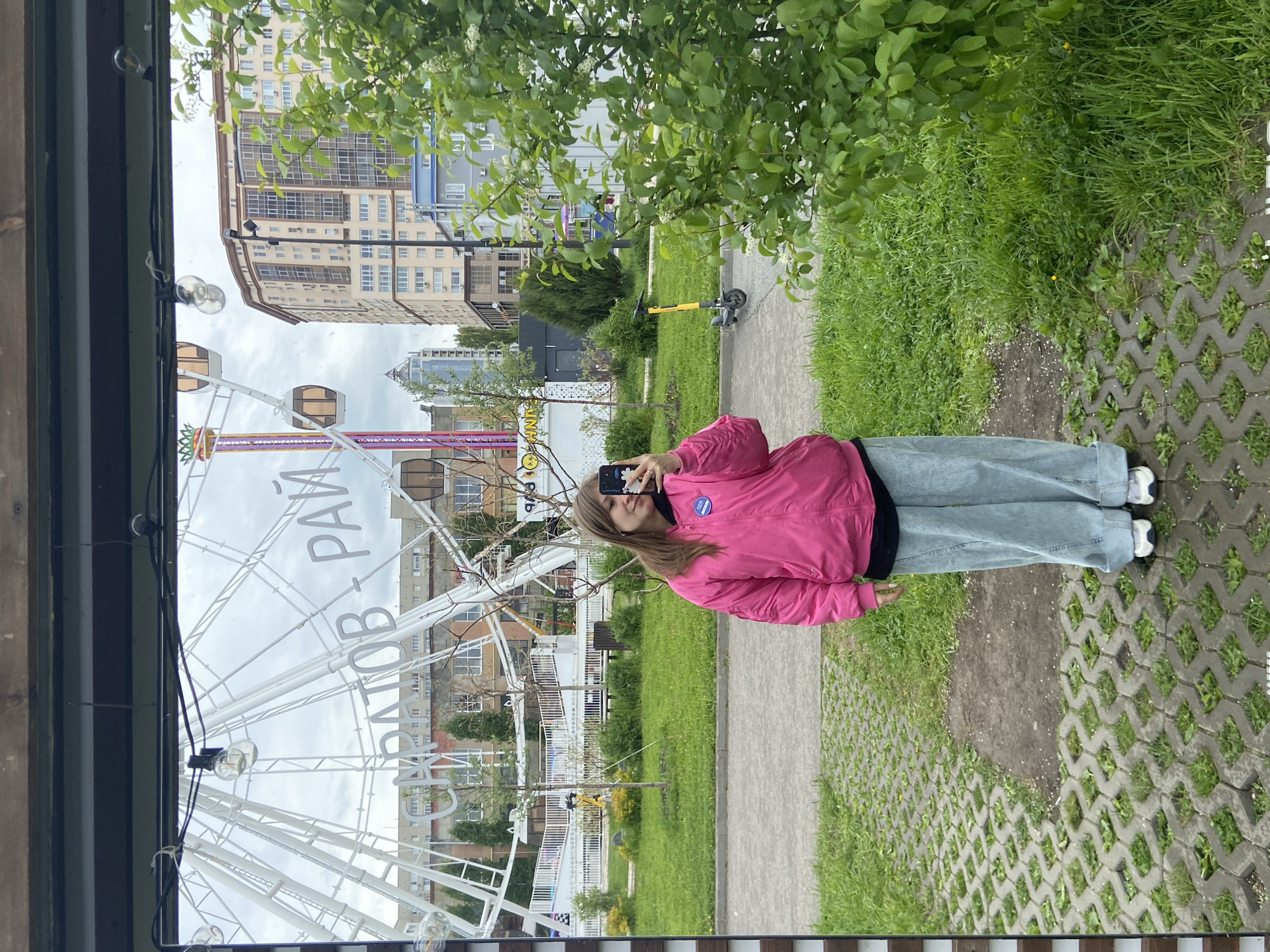 A woman in a pink jacket and light-wash jeans stands on a patterned concrete walkway, taking a selfie with her smartphone. Behind her is the Saratov Ferris wheel, identified by the Cyrillic text “САРАТОВ РАЙ” on its structure. The Ferris wheel is located in a park setting with trees and greenery. In the background, there are multi-story buildings, including a tall modern office or apartment building with numerous windows. A yellow electric scooter is parked in the distance. The lighting suggests it is daytime. The photo appears to have been taken from within a covered structure, with visible lights and supports framing the view.