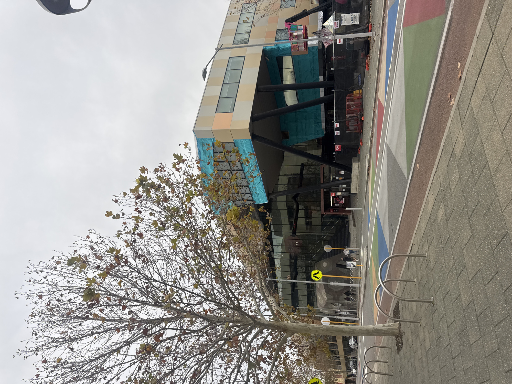 The image shows the Perth Cultural Centre in Perth, Western Australia, on an overcast day. A large tree with sparse autumn foliage stands in the foreground on the left, its branches bare. To the right, a modern building with a multi-colored facade (peach, beige, and white) is visible. The building appears to be under construction, with turquoise sheeting covering parts of its exterior and support beams. A construction lift with a pink cage is positioned on the right side of the building. A paved sidewalk with bike racks runs along the bottom of the image. The street in front of the building features a colorful, geometric pedestrian crossing. Several people are walking on the sidewalk in the middle ground, and cars are visible on the street in the background. Yellow pedestrian crossing signs with black arrows are placed along the sidewalk.