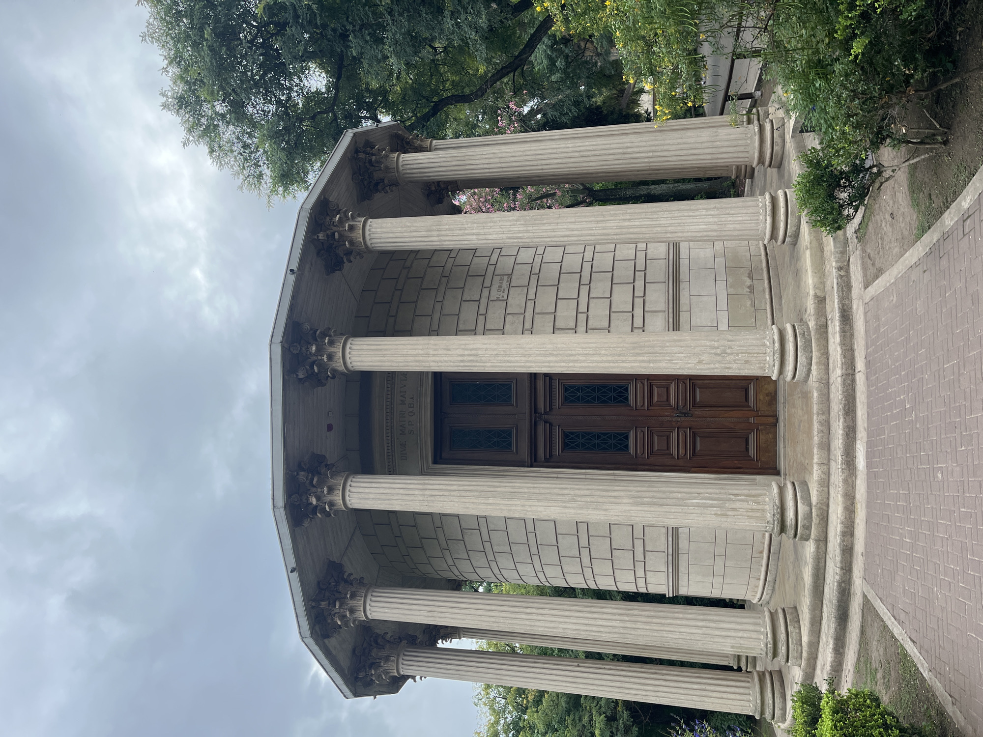 An octagonal classical-style mausoleum, identified as the Monumento a la Madre, is centrally positioned within the Recoleta Cemetery in the Recoleta neighborhood of Buenos Aires, Argentina. The structure is composed of light-colored stone, featuring eight prominent fluted columns with ornate Corinthian capitals supporting a flat, overhanging roof. Its walls are constructed from rectangular stone blocks. A pair of dark brown double wooden doors, detailed with square panels and rectangular glass inserts with diagonal crosshatch patterns, constitutes the primary entrance. Above these doors, the inscription "DIVE MATRI MATUTAE S.P.Q.B.A." is visible. An additional, smaller rectangular plaque or inscription is affixed to the left wall, its text illegible from this distance. The mausoleum rests on a multi-tiered, circular stone base with a series of shallow steps. A brick pathway, laid in a herringbone pattern, leads directly to the monument. The surrounding environment includes trimmed green shrubs flanking the pathway and dense green trees, some exhibiting pink and yellow blossoms, behind and to the sides of the structure. The sky overhead is uniformly overcast and gray. No individuals or animals are present within the captured scene.