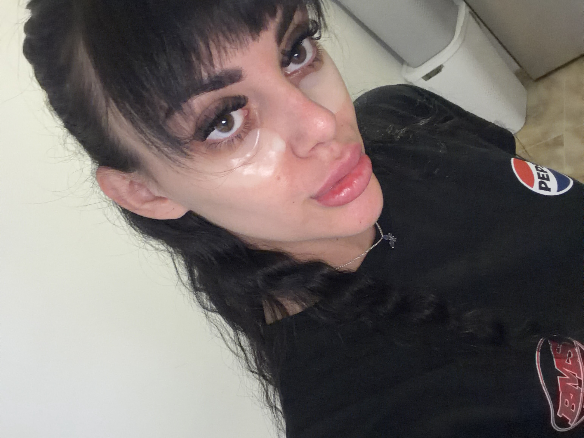The image shows a close-up, overhead, selfie-style shot of a young adult with dark hair and bangs, wearing a black t-shirt with a Pepsi logo on the right side of the chest and another logo on the left side. They have eye patches under their eyes, full lips, and are looking towards the camera. The background is a plain, light-colored wall, and to the right, a portion of a white trash can is visible. The image does not provide any specific details or context that would allow for identification of its location within the city of Plovdiv, Bulgaria.
