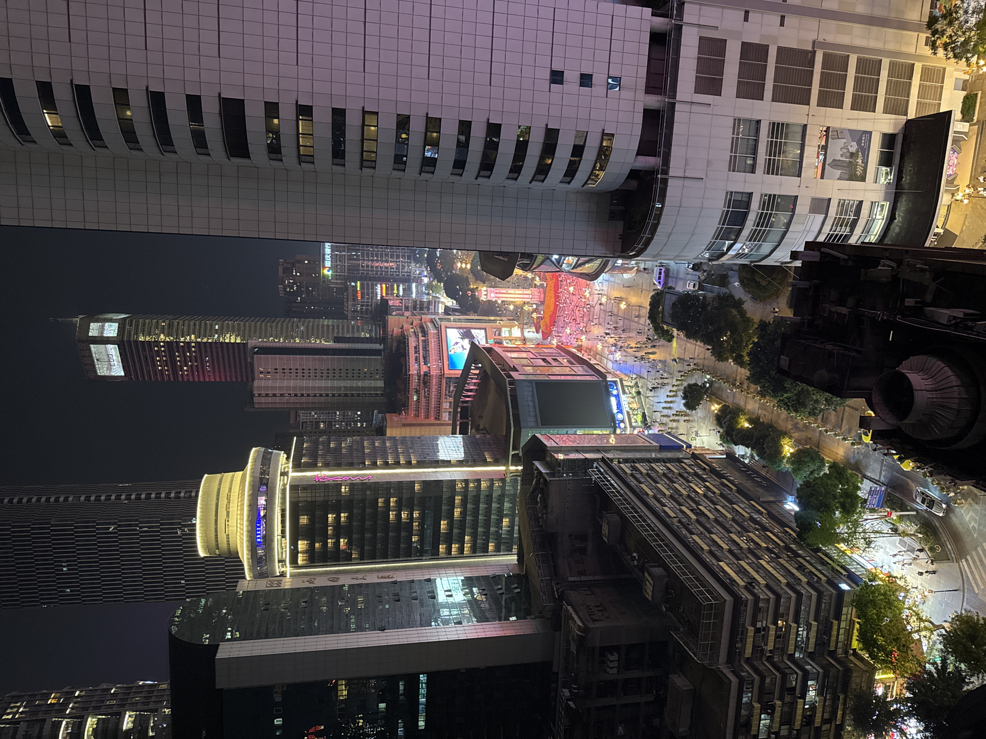 Captured from an elevated perspective in Yuzhong District, Chongqing, China, the image showcases a densely populated urban landscape at night. Illuminated skyscrapers and high-rise buildings dominate the skyline. Streets are filled with people, indicative of an active nightlife scene. The scene is brightly lit with streetlights and building illuminations. The presence of trees and foliage breaks the monotony of the concrete jungle. An aerial perspective offers a comprehensive view of the urban environment.