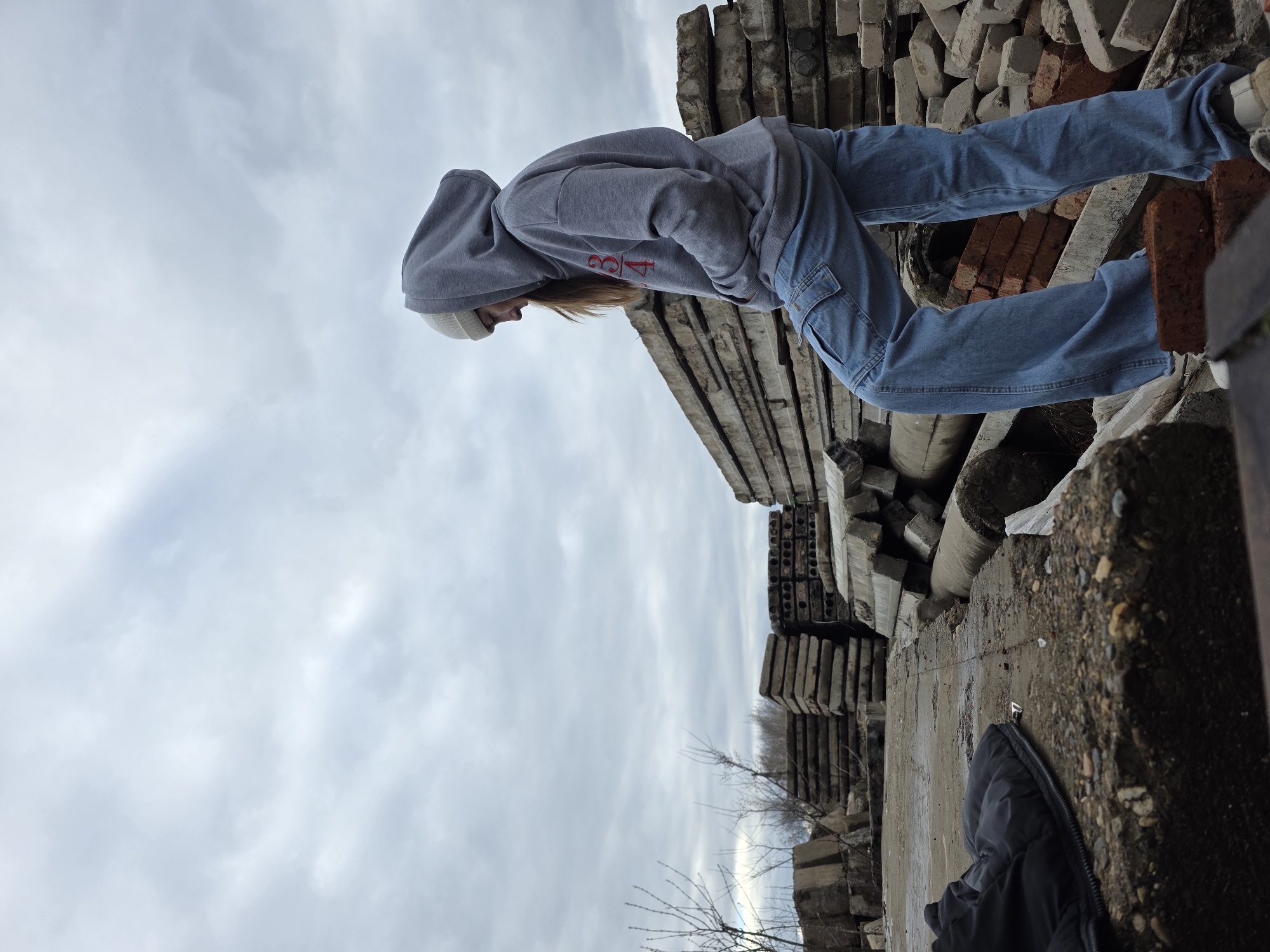 An outdoor, eye-level shot captures a person standing amidst stacks of construction materials under an overcast sky. The person, positioned towards the right side of the frame, is seen from the side, leaning forward and looking downwards. They are wearing a grey hooded sweatshirt with red text "3/4" or "3|4" visible on the front, blue jeans with cargo pockets, a light-colored beanie, and light-colored sneakers.
The setting appears to be an industrial area, construction site, or a demolition zone. In the background and around the person, there are numerous stacks of rectangular concrete slabs, some with visible hollow cores, and piles of red bricks. The ground is rough concrete or earth with pebbles and debris. Sparse, bare tree branches are visible in the distance against the light grey, cloudy sky. A black puffer jacket is lying on the ground in the bottom right corner of the image.
The weather is cloudy, suggesting a cool or cold day, possibly late fall or winter given the bare trees. The time of day is daytime, but the heavy cloud cover prevents bright sunlight. The person's posture suggests they are observing something on the ground below them or are in a thoughtful state.