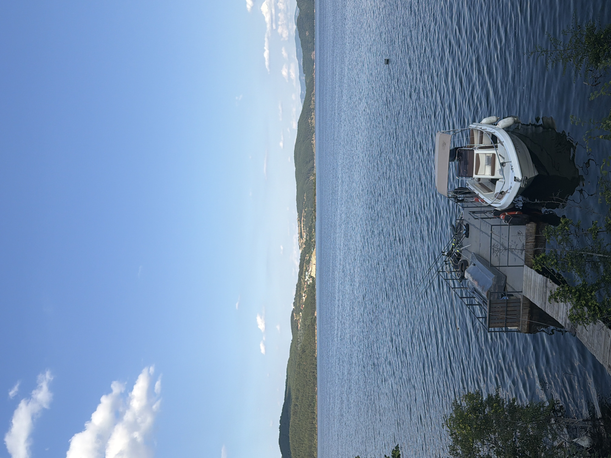 The image depicts a serene lake scene with a clear blue sky and scattered white clouds. In the foreground, a wooden dock extends into the calm, blue water. A small white motorboat with a tan canopy is moored to the dock, alongside other items such as life preservers and fishing rods. The water is rippled, suggesting a gentle breeze. In the background, a lush green, tree-covered hillside rises from the water's edge, dotted with houses and buildings, indicating a settlement. Further in the distance, more rolling hills are visible, creating a picturesque landscape. The overall impression is one of tranquility and natural beauty, likely a popular spot for recreation and relaxation in Zvanika, Bulgaria.
