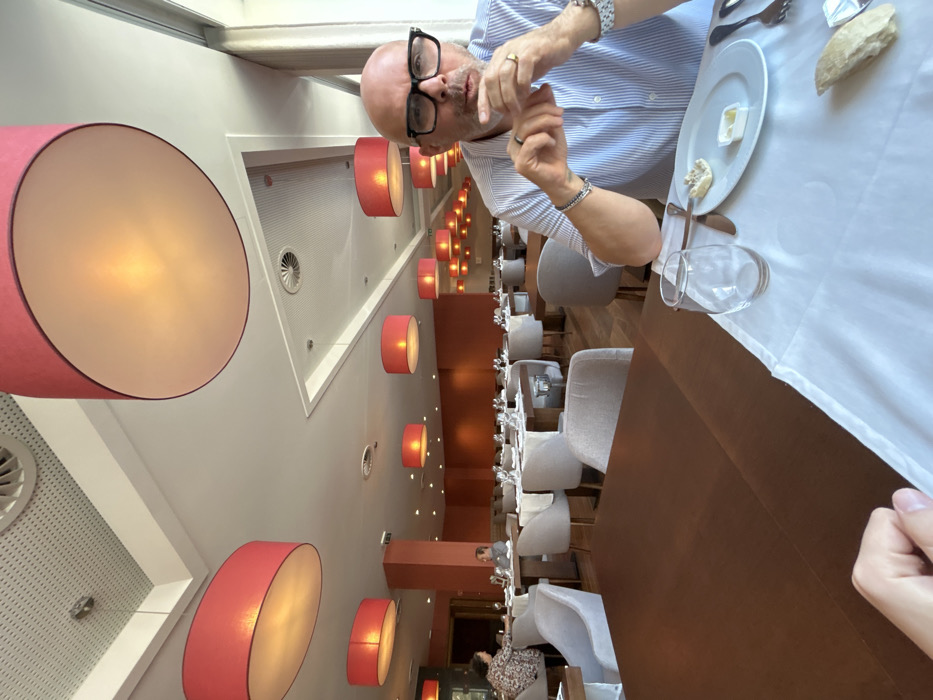 The image displays the interior of a restaurant, identified as being within Hotel Casino Chaves in Chaves, Portugal. In the foreground, a bald male with dark-rimmed glasses, wearing a light blue striped button-up shirt, is seated at a table on the right. His hands are clasped near his mouth, and he wears a gold-colored ring on his right ring finger, multiple bracelets on his left wrist, and a watch on his right wrist. On the white tablecloth in front of him are an empty stemless wine glass, silverware (fork, knife, spoon), a white plate containing a small piece of bread and a plastic container of butter, and a larger piece of bread. The photographer's left hand rests on the dark brown wooden table surface in the bottom left corner. The dining room is spacious, featuring numerous dark brown wooden tables, many set with white tablecloths, and light beige upholstered dining chairs. The white ceiling incorporates perforated acoustic panels and ventilation grilles, with multiple large, circular, red-orange fabric lampshades suspended, casting a warm light. The walls are painted an orange-tan color. Large windows are visible along the right side of the room, admitting natural light. In the mid-ground and background, other patrons are visible seated at tables, including a woman with curly hair and a patterned top on the far left, and a man seated alone further back.