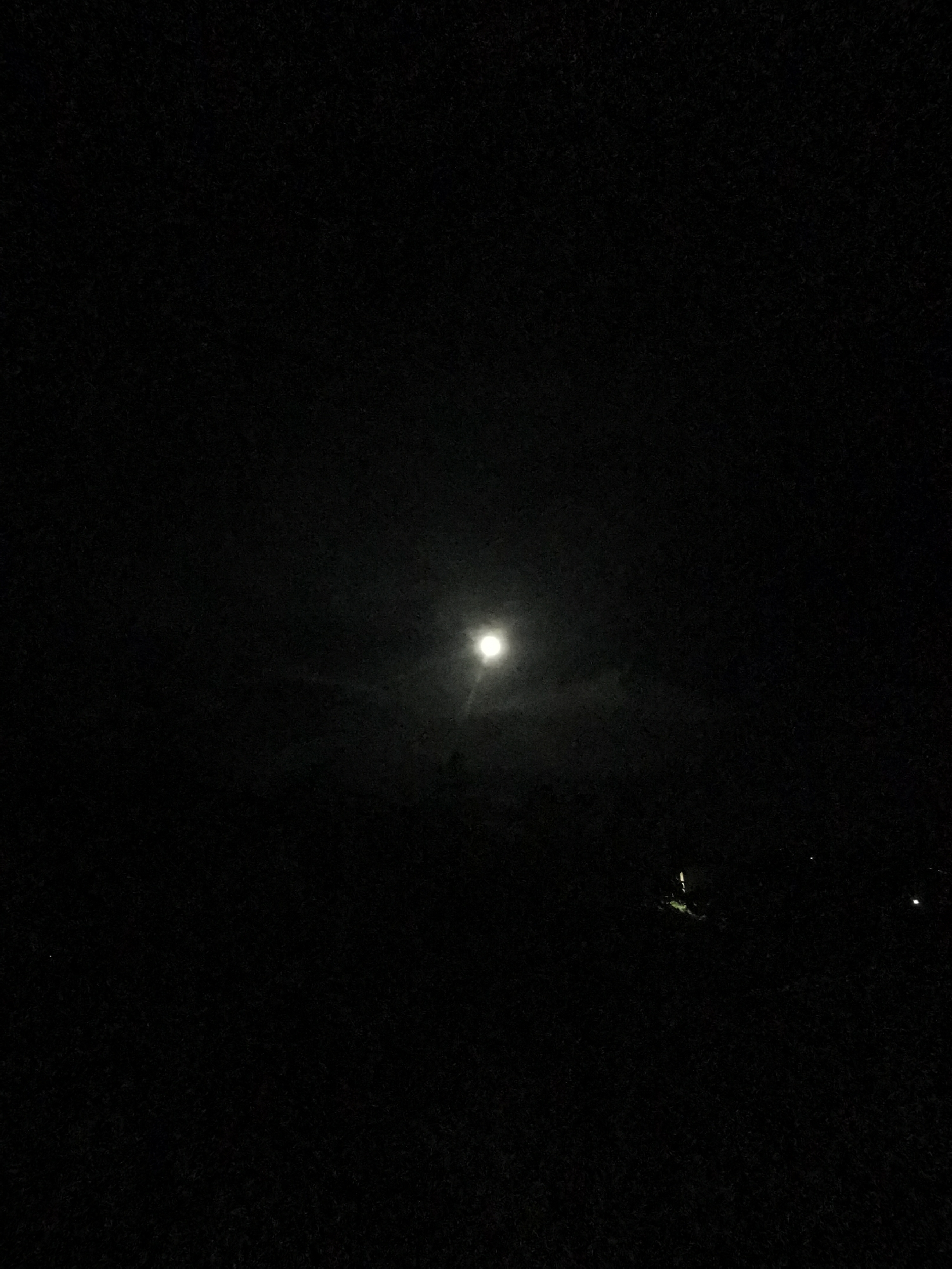 The image depicts a night scene in Ilindentsi, Bulgaria. The moon is visible in the center of the frame, with some surrounding cloud cover. The ground appears dark, and some faint lights are visible in the distance. A small cluster of brighter lights is also discernible on the lower right side of the image, perhaps from a building or streetlights. Overall, the image is quite dark and details are difficult to discern.