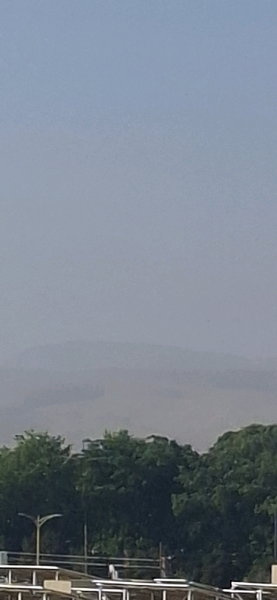 The image captures a hazy, light blue sky dominating the upper two-thirds of the frame. Below the sky, indistinct layers of distant hills are visible, shrouded in atmospheric haze, their outlines softened and muted. In the foreground, a dense line of green trees stretches across the lower portion of the image. Above the trees, the tops of some low-rise buildings are peeking through, suggesting an urban or semi-urban environment. A street lamp with a distinctive curved arm is also visible to the left of the frame, emerging from behind the trees. The overall impression is one of a slightly overcast or dusty day, obscuring distant details and lending a soft, diffused light to the scene.