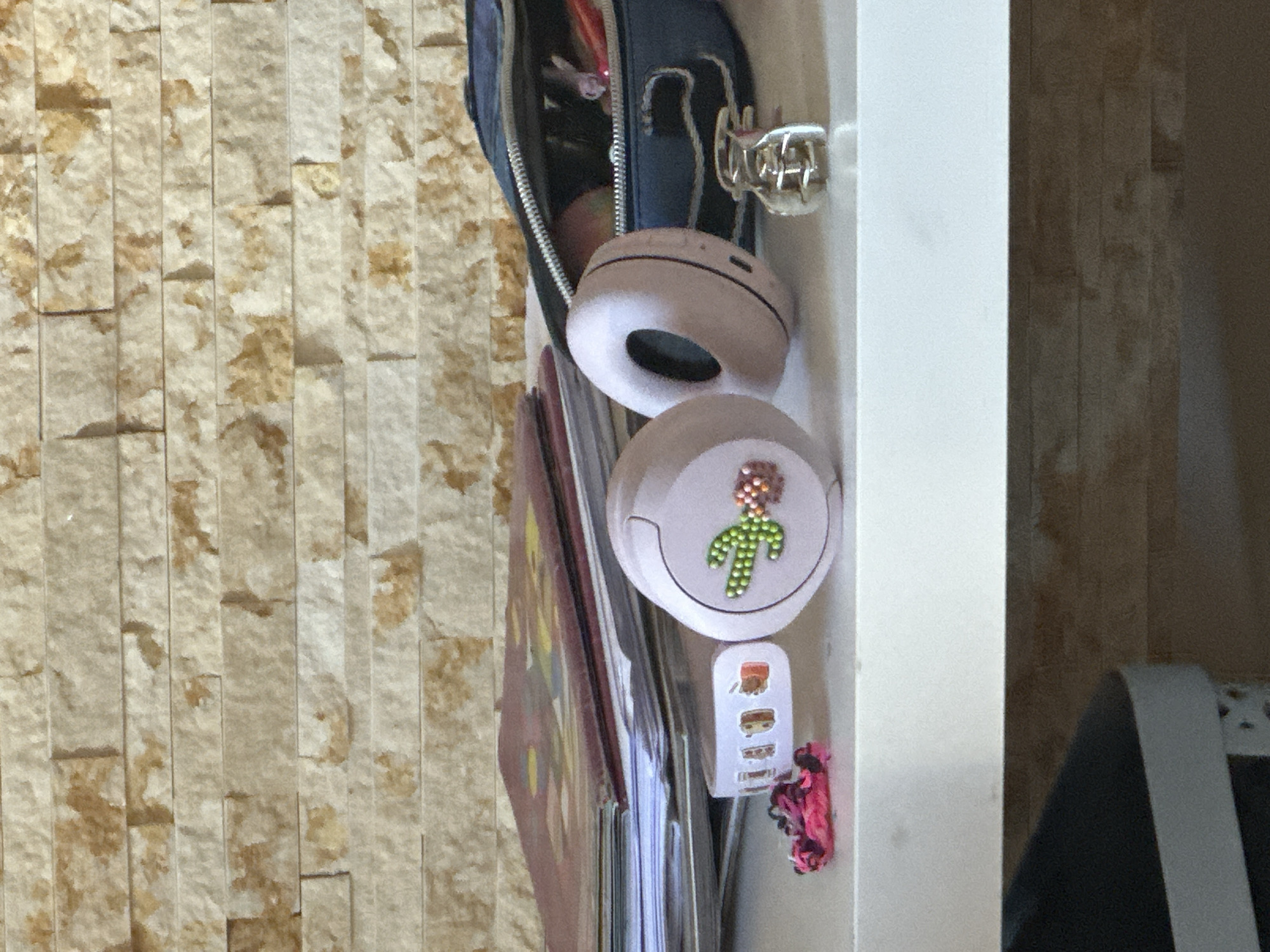 A close-up shot, captured at a slightly high angle, presents a collection of objects on a white shelf, set against a textured stone-patterned wall. In the foreground, two light pink wireless headphones are visible. The left headphone has a sparkling green cactus design on its ear cup, accented with small pink and orange rhinestones. To the left of the headphones, a bright pink, knotted hair tie rests on the shelf. Further left, a white sticker band with small illustrations of fruit and other designs is also present. Behind the headphones, a stack of papers and books is arranged. A dark blue zippered pencil case sits to the right of the headphones, partially open, with a glimpse of colorful pens and possibly an apple inside. A small, silver metal hair claw clip is positioned to the right of the pencil case. The overall scene suggests a desk or study area. The background wall features a pattern of horizontally stacked rectangular stones in various shades of beige and tan, creating a naturalistic texture. The lower portion of the image reveals a dark grey surface with a white stripe, possibly the edge of another shelf or furniture item. The image is well-lit, with the light source appearing to be from the front and slightly to the left. No people are present in the scene.