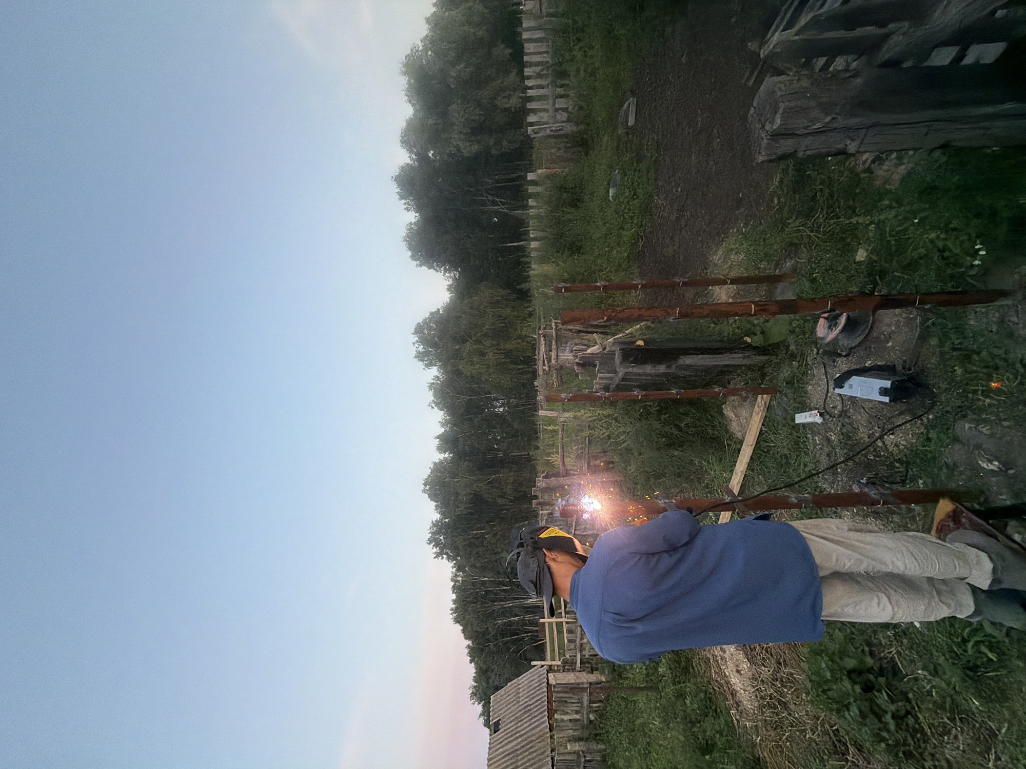In the village of Kuseevo, Russia, a person is welding metal posts in an outdoor setting at dusk. The individual, wearing a blue shirt, beige pants, a dark baseball cap, and a welding helmet with yellow protective lenses, is standing on a small platform. Sparks are visibly emanating from the welding torch as it is used on a metal pole. The environment is rural, with a wooden fence, overgrown grass, and trees in the background. A wooden structure resembling a shed or outbuilding is visible to the left. The sky is a soft blue, transitioning to a lighter hue near the horizon. A welding machine and cables are on the ground nearby.