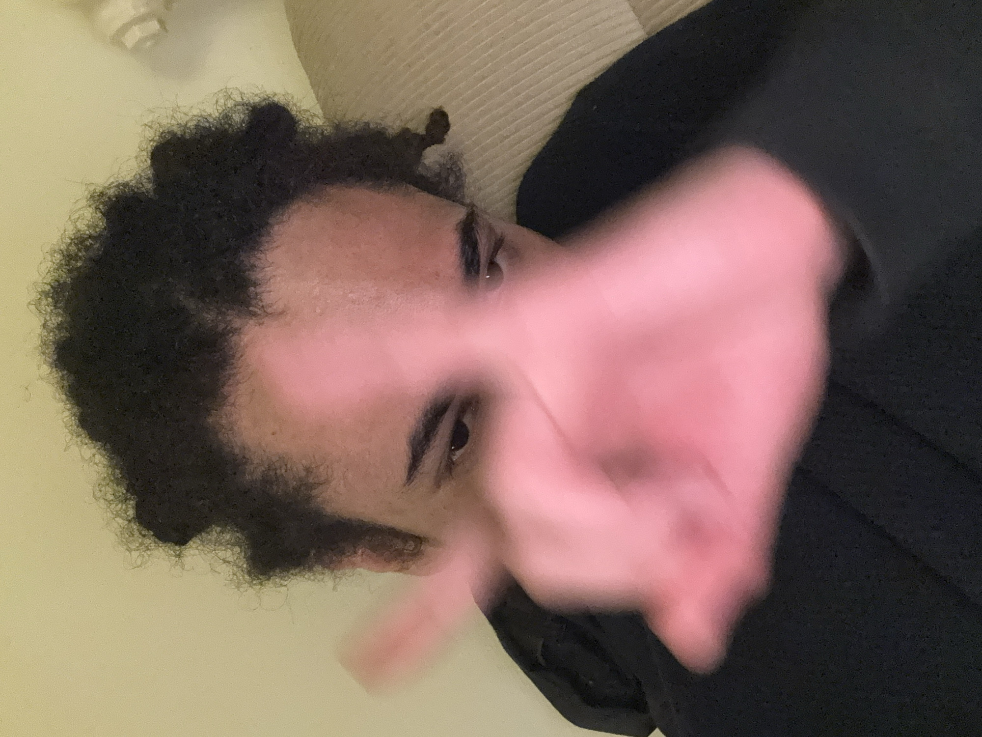 The image displays a close-up view of an individual, likely an adult, situated indoors within the city of Paris, France. The subject's face is partially obscured by a highly blurred, reddish-hued hand positioned prominently in the foreground, suggesting rapid movement during image capture. Visible facial features include dark, curly hair, dark eyes looking downwards and to the viewer's right, and a small piercing near the inner corner of the left eye. The individual is wearing a dark, possibly black, garment that covers the shoulders and torso. The background consists of a light, pale yellow wall on the left and a textured, light beige or cream surface, possibly upholstery or bedding, on the right. A small, indistinct white fixture is partially visible in the upper right background. The scene is illuminated by soft, warm artificial light.