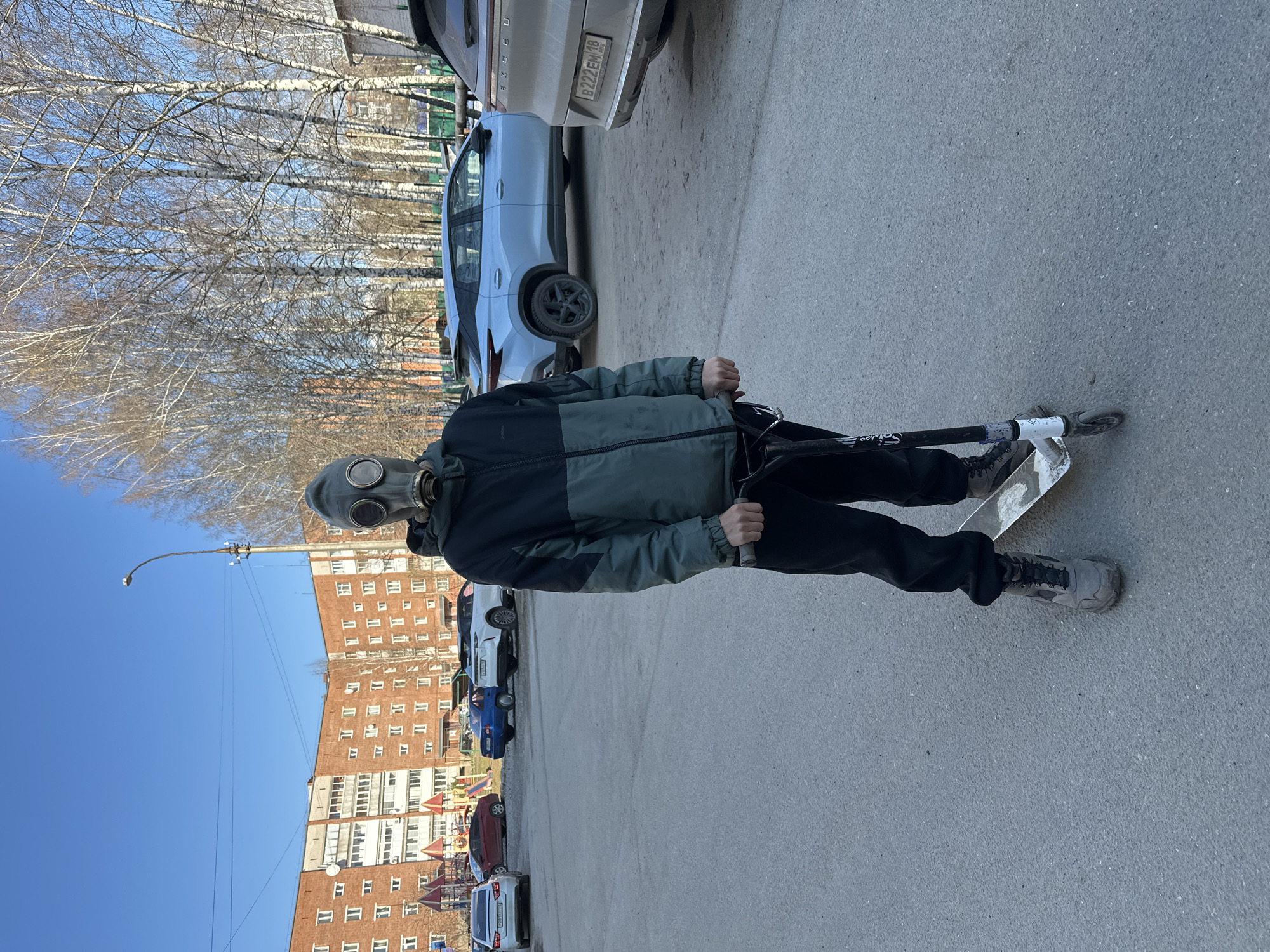 A person is depicted outdoors, standing on a paved surface, possibly a street or parking lot. The individual wears a two-tone dark green and black jacket, dark pants, and grey athletic shoes. Most notably, their face is entirely covered by a grey gas mask with prominent eye lenses and a central filter. They are holding the handlebars of a white and black kick scooter, with one foot on the scooter's deck and the other on the ground.
In the background, a row of tall, slender trees with bare, white-barked branches, likely birches, stands beneath a clear blue sky. Behind these trees and cars, a large, multi-story red-brick apartment building with numerous windows and some balconies is visible. A lamppost rises on the left side of the frame, near the building. Several vehicles are parked along the side of the road or in parking spots. One light-colored SUV, possibly an Exeed, shows a Russian license plate that reads "B222EM 18". Another license plate, partially visible on a white car, appears to be "O266AA 18". The scene suggests a bright, sunny day, likely in late winter or early spring given the bare trees. A children's playground structure can be partially seen near the base of the apartment building.