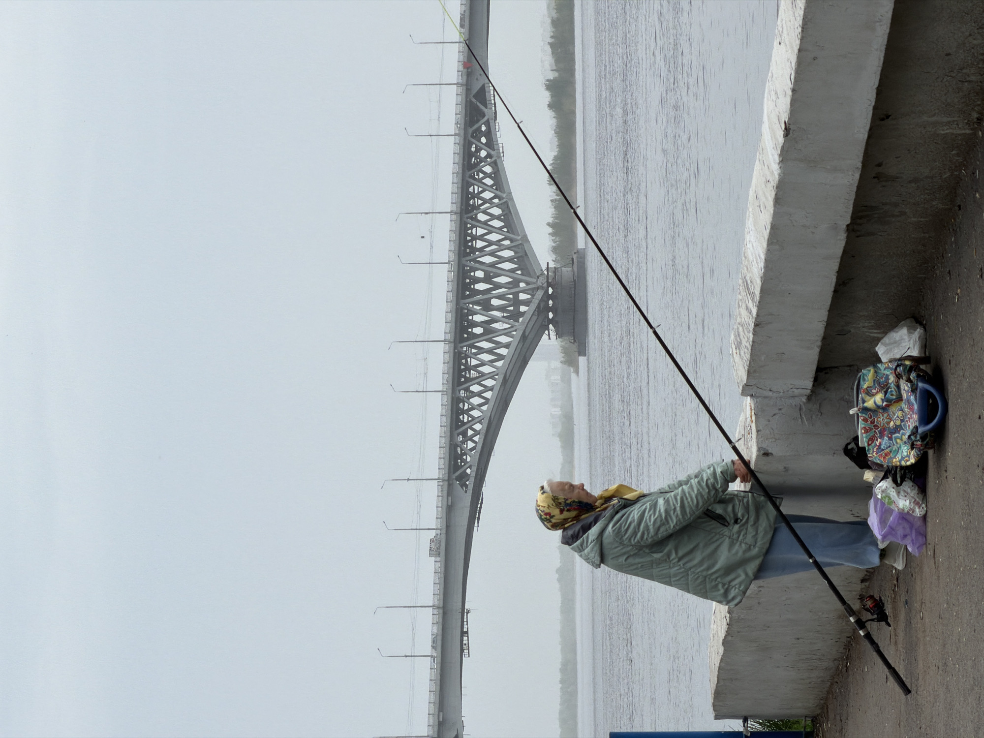 A horizontal medium shot depicts an older woman fishing off a concrete embankment with a bridge visible in the background, taken in the city of Saratov, Russia.
The woman stands facing right, wearing a patterned headscarf, a light green quilted jacket, and light blue pants. She holds a long fishing rod that extends over the water. To her right, against the concrete wall, is a colorful backpack and some white plastic bags. The concrete embankment is light gray and shows signs of wear.
Behind the woman and the embankment is a body of water, most likely the Volga River, with a steel bridge visible in the background. The bridge has a distinctive arched structure. The sky is overcast and gray.