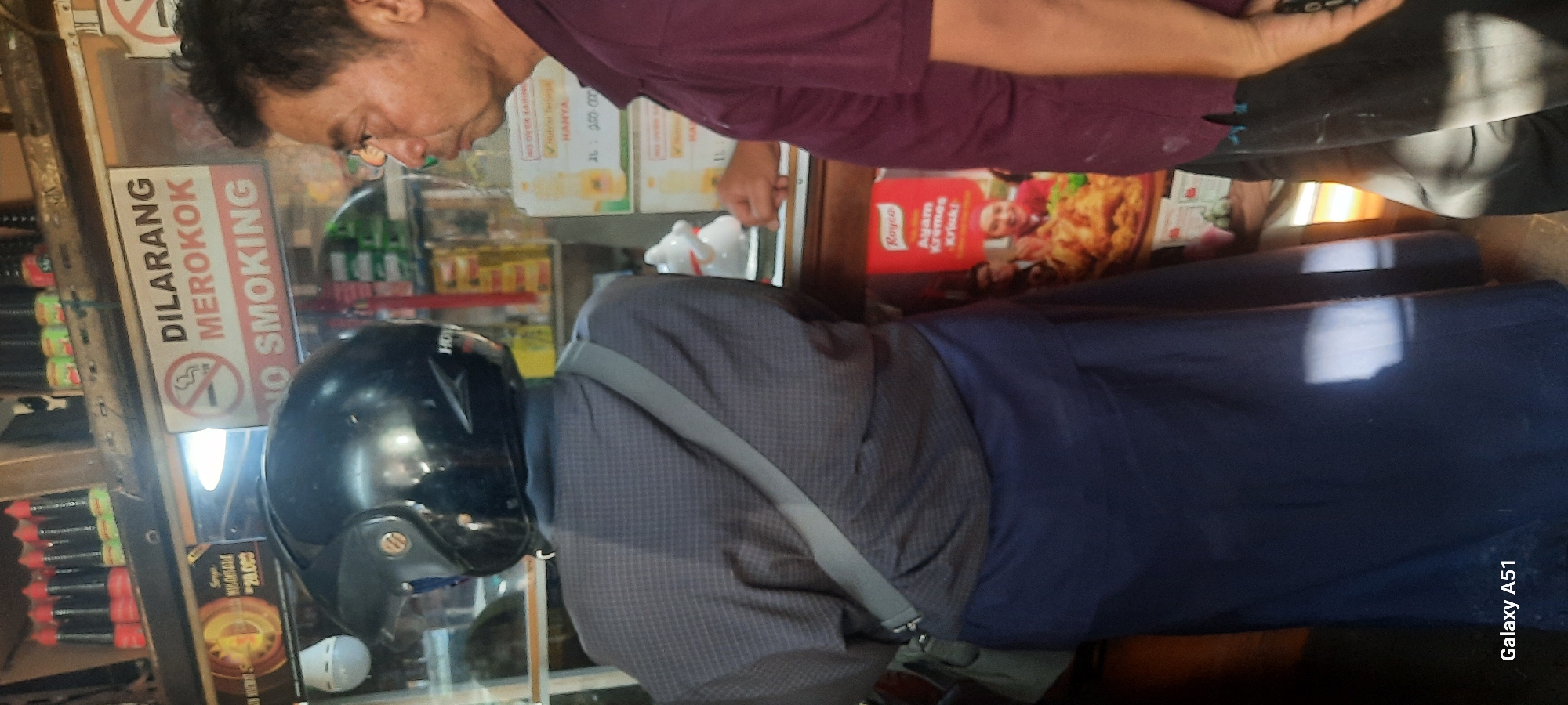 In a dimly lit convenience store in Beru, Indonesia, a person wearing a dark checkered jacket and a blue skirt stands with their back to the camera, facing shelves stocked with various goods. They have a black helmet on their head and a grey strap across their chest. To their right, a man in a maroon shirt and dark pants looks towards the camera. Behind them, a "NO SMOKING" sign is prominently displayed on the wall. The shelves are filled with bottles of condiments and other packaged items. A poster advertising fried chicken is visible on the right side of the image. The store appears to be a small, local establishment.