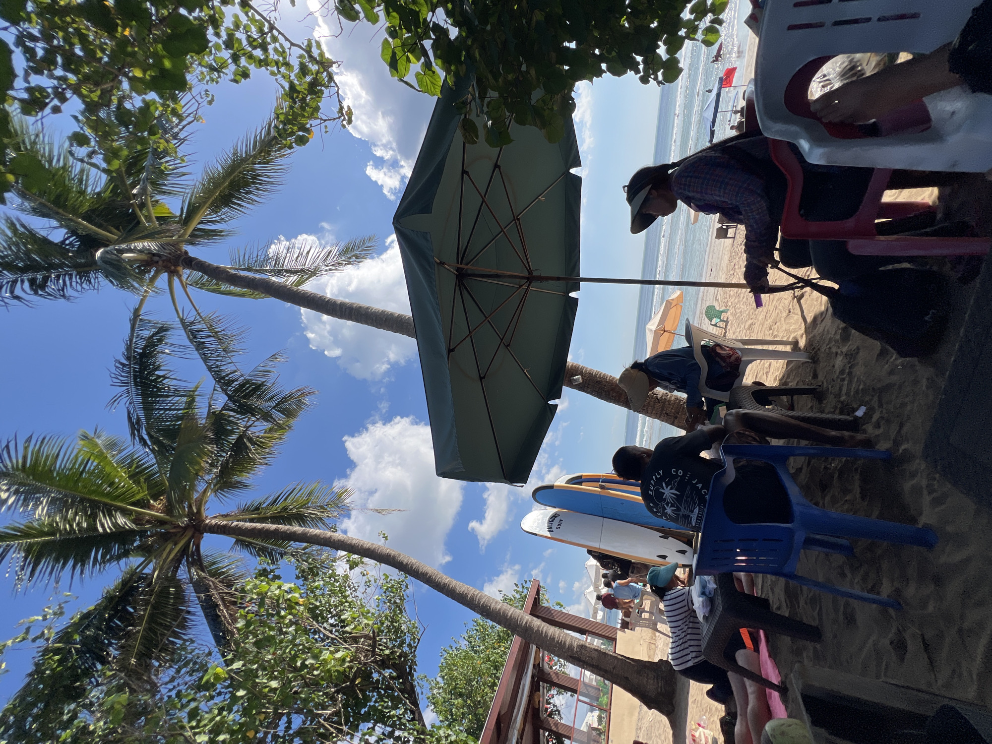 The image depicts a beach scene in Kuta, Indonesia, under a bright, partly cloudy sky. Several palm trees are visible, with lush green foliage. In the foreground, people are seated at tables on the sandy beach. A large, dark green umbrella provides shade. Two surfboards are leaning against each other, with one displaying the text "BALIGURU SURF." A person in a blue shirt and a person in a black shirt with a palm tree graphic are seated at a table. Another person wearing a hat and a plaid shirt is seated in a red chair. Beach chairs and tables are scattered on the sand. In the background, the ocean can be seen with gentle waves, and a few people are visible in the water and on the beach. A building with a red roof is situated further down the beach. The overall atmosphere suggests a relaxed beachside cafe or seating area.