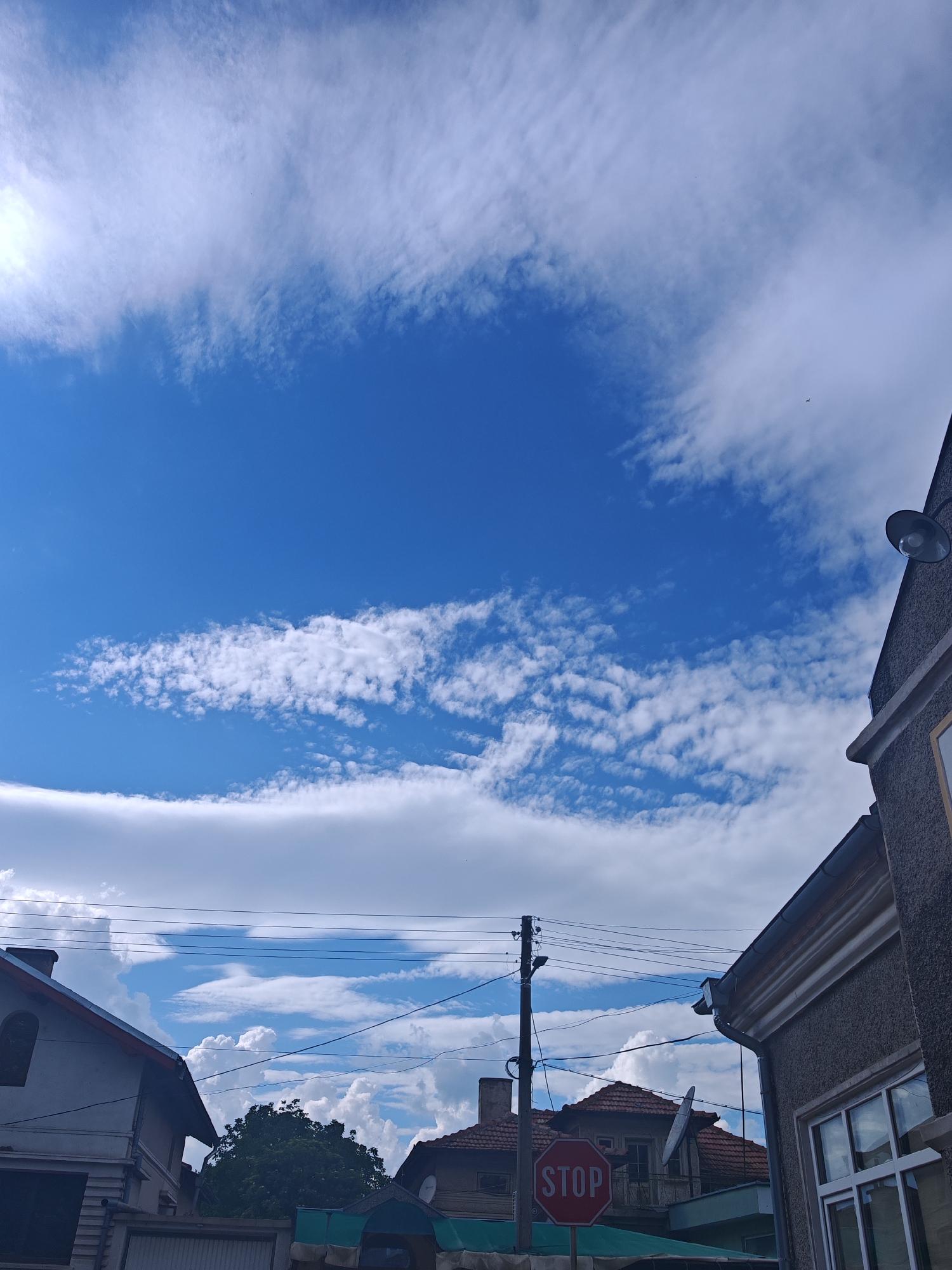 The scene is an upward-angled view captured in Tsarev Brod, Bulgaria, primarily depicting a dynamic sky and the upper sections of residential structures. The sky occupies approximately two-thirds of the image, showcasing deep blue areas interspersed with three distinct cloud layers: diffuse, high-altitude white clouds dominate the upper left and right, a clear band of blue sky stretches horizontally across the mid-upper section, and a more defined, broken band of white altocumulus clouds extends horizontally below the clear blue. A minuscule, dark, winged object, likely an aircraft or bird, is visible against the clouds in the upper right quadrant.
The lower third of the image frames portions of buildings and utility infrastructure. On the far left, a section of a light-colored house with a dark-tiled pitched roof and an upper-level window is partially visible. In the middle ground, a dark utility pole rises, supporting multiple black overhead power lines that crisscross the frame horizontally, extending between buildings. A red octagonal STOP sign with white lettering is prominently positioned in the lower-middle, mounted on a pole and partially obscuring the front of a background building. This background building features a reddish-brown tiled, pitched roof, a visible chimney, and a circular satellite dish affixed to its facade. On the far right, a dark grey, textured building facade is present, revealing a white-framed window on the lower right and a dark, wall-mounted outdoor lamp near its top edge. Dense green tree foliage is visible in the background, positioned between the structures. No people are visible within the frame.