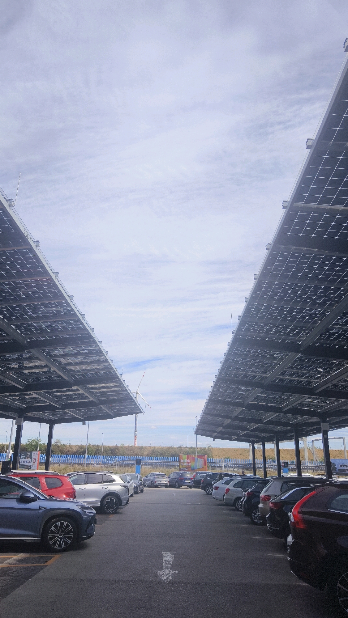 The image shows a parking lot with two rows of solar panel canopies on either side, creating a pathway for cars. The parking lot is filled with various cars parked in designated spots. In the background, a wind turbine is visible, along with a fence and some vegetation. The sky is cloudy with patches of blue. The scene appears to be in an open area, possibly a commercial or industrial zone. The presence of the solar canopies suggests a focus on renewable energy. The image does not contain any people or specific details that would allow for pinpointing a precise location within Vösendorf, Austria.