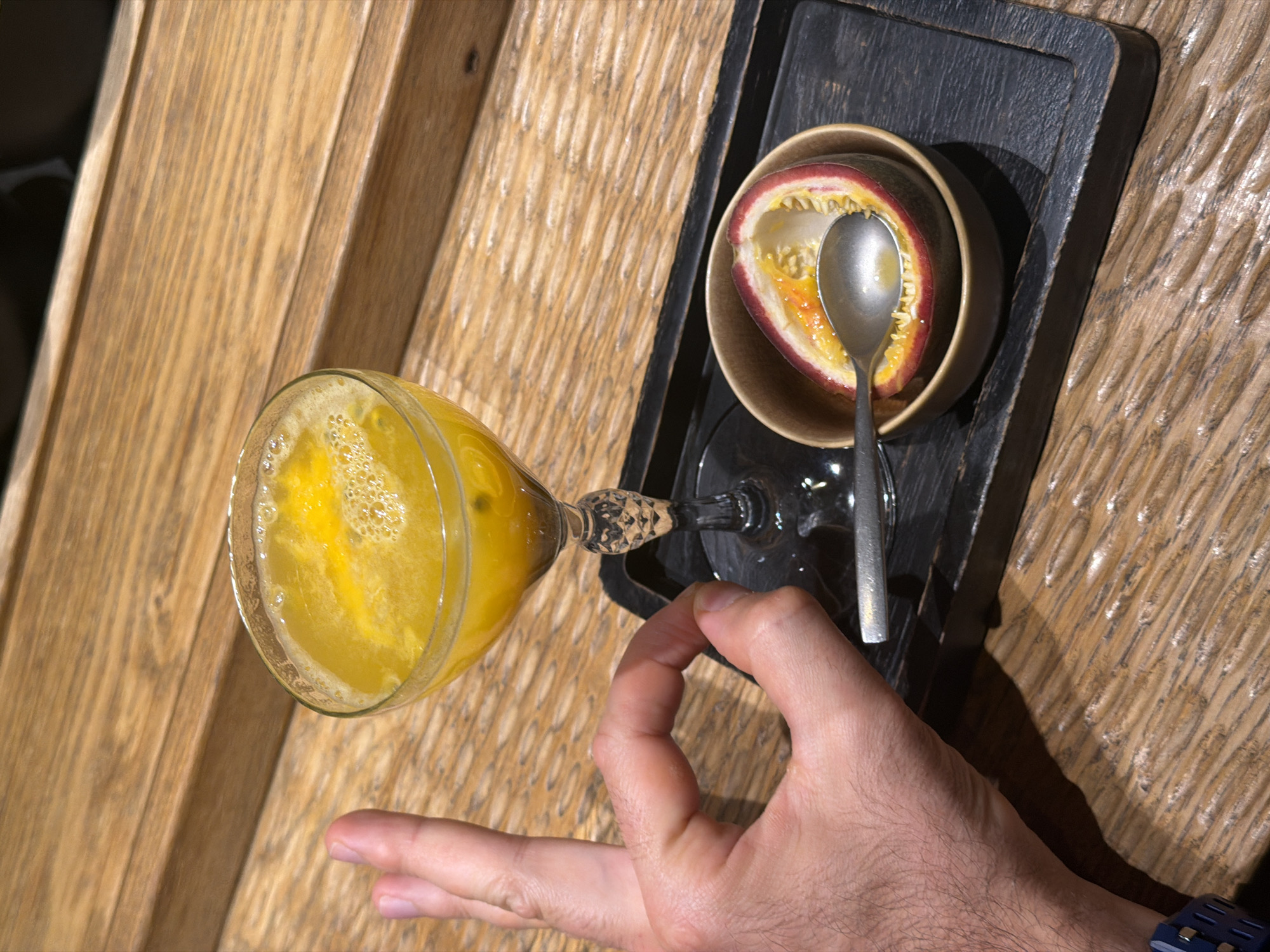 An overhead shot captures a hand reaching towards a stemmed glass filled with a vibrant yellow, frothy beverage, likely a juice or cocktail, against a backdrop of a wooden surface. The hand, exhibiting an "OK" gesture, has a blue watch band visible on its wrist.
The glass has a decorative, faceted stem and is positioned on the left side of the frame. To its right, on a dark, textured rectangular tray, sits a small, light brown bowl containing a halved passion fruit. The fruit's bright orange-yellow pulp and black seeds are exposed, and a silver spoon rests within it, ready for consumption.
The dominant background is a light-colored wooden table or paneling, featuring a distinctive vertical grain pattern. The overall presentation suggests a refined dining experience or a refreshment served in an indoor setting, possibly in Krasnoyarsk, Russia. No visible text is present.
