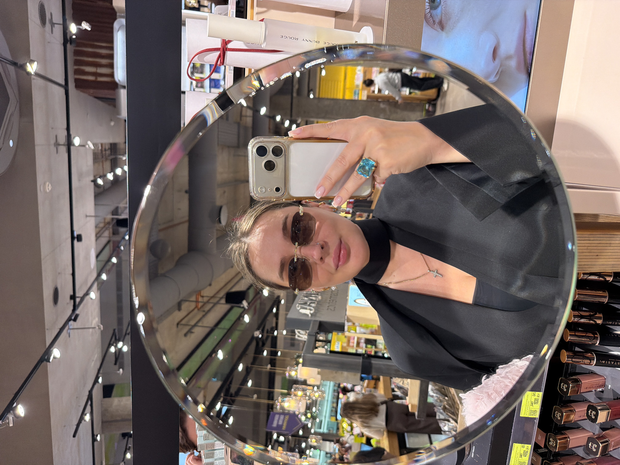 A person is taking a selfie in a round mirror within a retail setting. The person is wearing sunglasses, a black top, and a ring with a large, square, blue stone. They are holding a smartphone with a gold case in their right hand. The mirror reflects the interior of what appears to be a store, with shelves of cosmetic products visible. Reflected in the mirror are overhead lights, and in the background, other shoppers are visible. The retail setting is located in Moscow, Russia.