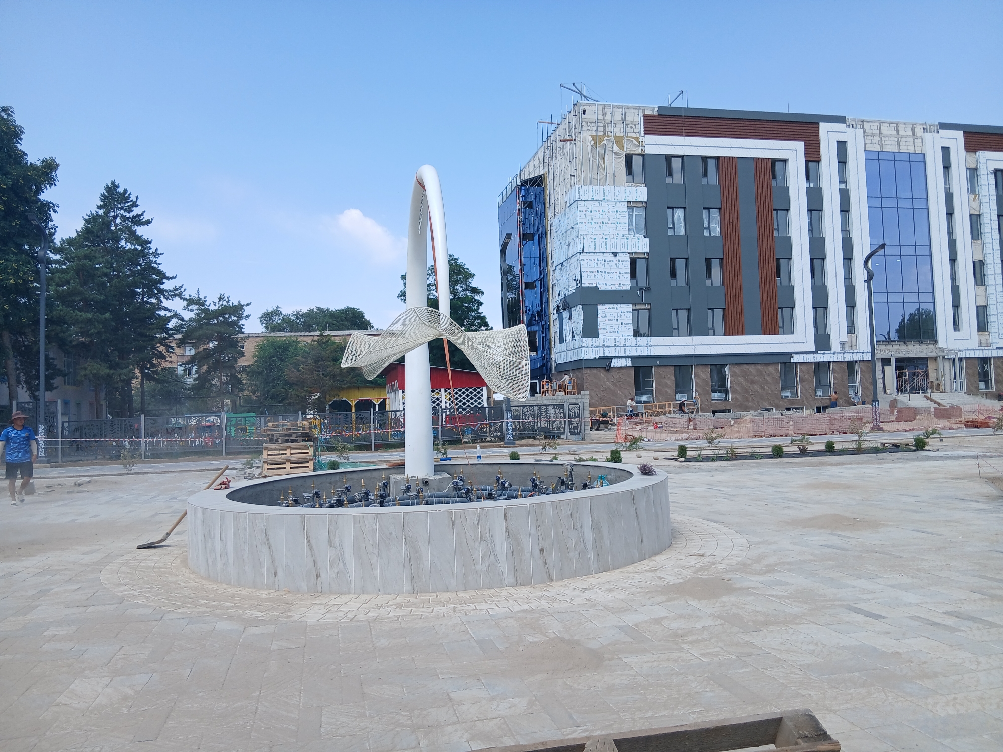 In the city of Talas, Kyrgyzstan, an outdoor scene depicts a public space under construction. In the foreground, a circular fountain with a grey, stone-like base is visible. The fountain appears to have multiple water spouts. A white, curved sculpture with a woven net-like element is positioned above the fountain. The ground surrounding the fountain is paved with light-colored rectangular tiles, with some areas showing loose sand and construction debris, including a broom and a shovel.

Behind the fountain, a large, multi-story building is under construction. The building has a modern design with grey, white, and brown sections, featuring numerous windows, some of which are covered with protective film. Large blue-tinted windows are also prominent on one facade. Portions of the building's exterior are covered with white insulation material, indicating ongoing work. Construction equipment and materials, such as stacks of bricks and wooden pallets, are scattered around the building.

To the left of the building, mature trees are present, providing a contrast to the ongoing construction. In the background, a fence separates the construction area from other parts of the city. A person wearing a blue t-shirt and shorts, and a hat, is walking away from the camera on the paved area to the left. The sky is clear and blue with a few wispy clouds. The overall scene suggests a developing urban environment, with public amenities and infrastructure being updated.