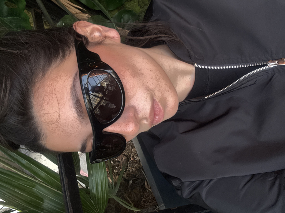 A person with dark hair and freckled skin wears black sunglasses and a black jacket. Part of a building's exterior is reflected in the sunglasses' lenses. Green foliage is visible in the background. The jacket has a silver zipper. The person's face is in focus, with a slight smile. The scene appears to be outdoors during the day.