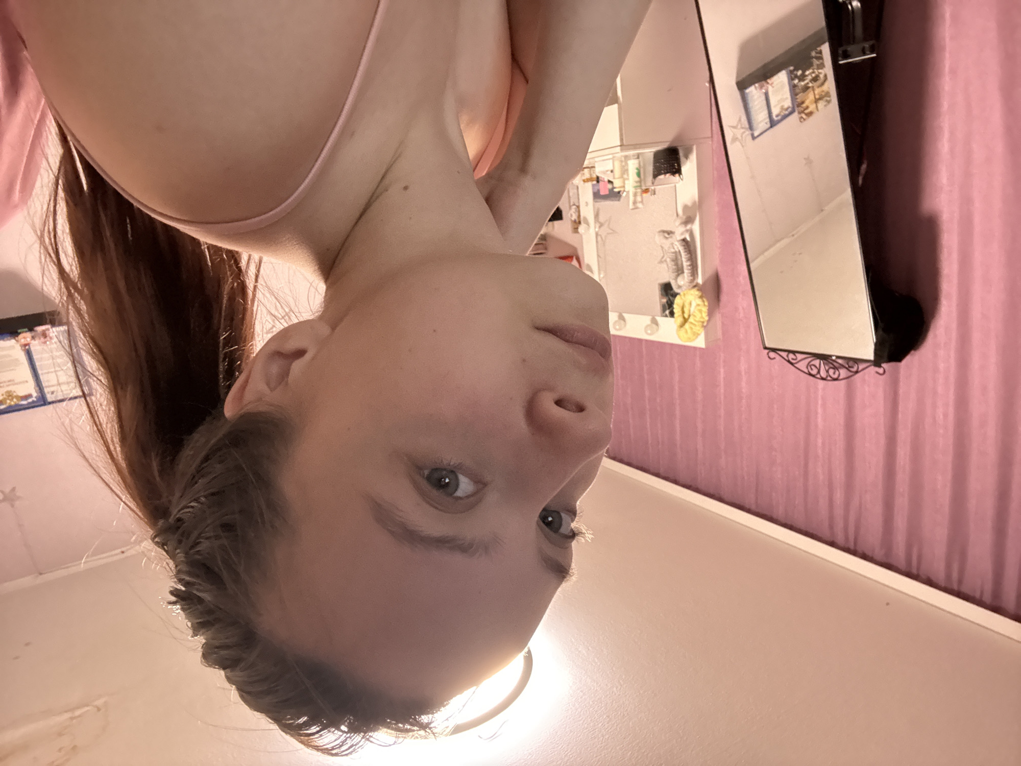 A close-up, upside-down perspective shot depicts a fair-skinned individual with light brown hair pulled back in a ponytail. The person is wearing a pink garment, potentially a tank top or camisole. Facial features, including eyes, nose, and mouth, are visible and slightly out of focus due to the close proximity. The background includes a light pink curtain, a vanity with a mirror reflecting various cosmetic products and decorations. A small bulletin board with papers and a framed object are also visible on the wall. The specific location in Прикамский, Russia cannot be determined from the provided image.