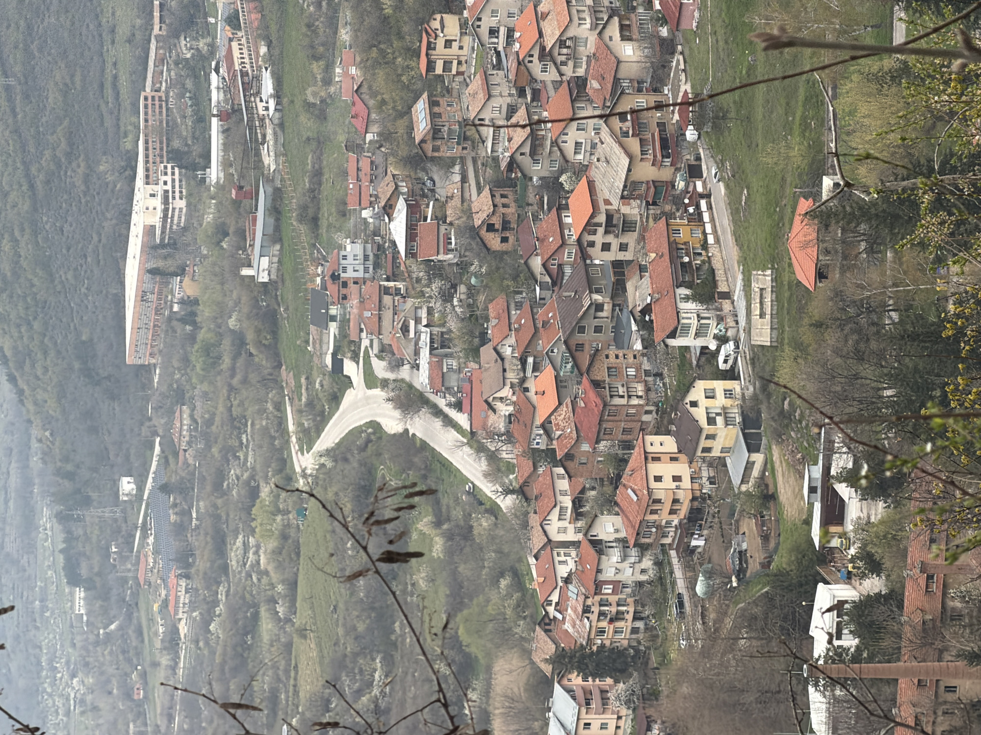 The image depicts a high-angle view of a residential area, characterized by densely packed houses with orange and brown tiled roofs, nestled amongst trees and greenery. A road winds through the landscape and the background shows hills and a mountainous region. The image appears to be taken in daylight, with an overexposed and slightly hazy atmosphere. Based on the description, the location is likely Bratsigovo, Bulgaria. The scene has a mix of residential buildings, foliage, and infrastructure, as one might observe from a vista point. There are no visible people or interactions in the captured perspective.