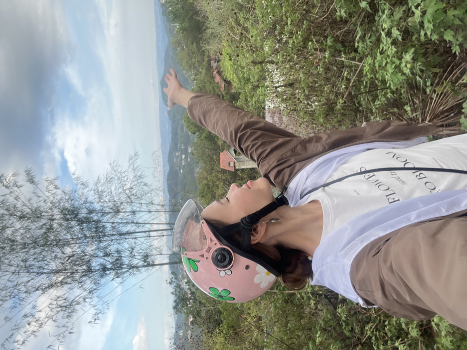 A young adult female is depicted outdoors at an elevated viewpoint in Da Lat, Vietnam, with a panoramic view of hills and residential areas in the background under a partly cloudy sky. She is seen in profile, facing right, with her eyes closed and a slight smile, suggesting contentment. She wears a light pink motor scooter helmet adorned with green four-leaf clovers and white daisy decals, a brown jacket over a white t-shirt with black text, and a silver-colored tear-drop earring on her visible right ear. Her right arm is extended upwards and slightly forward, with her palm open and facing the sky. Tall, slender bamboo stalks rise on the left side of the frame. The foreground and mid-ground are covered in various green foliage, with several houses featuring red roofs visible amidst the distant hills.