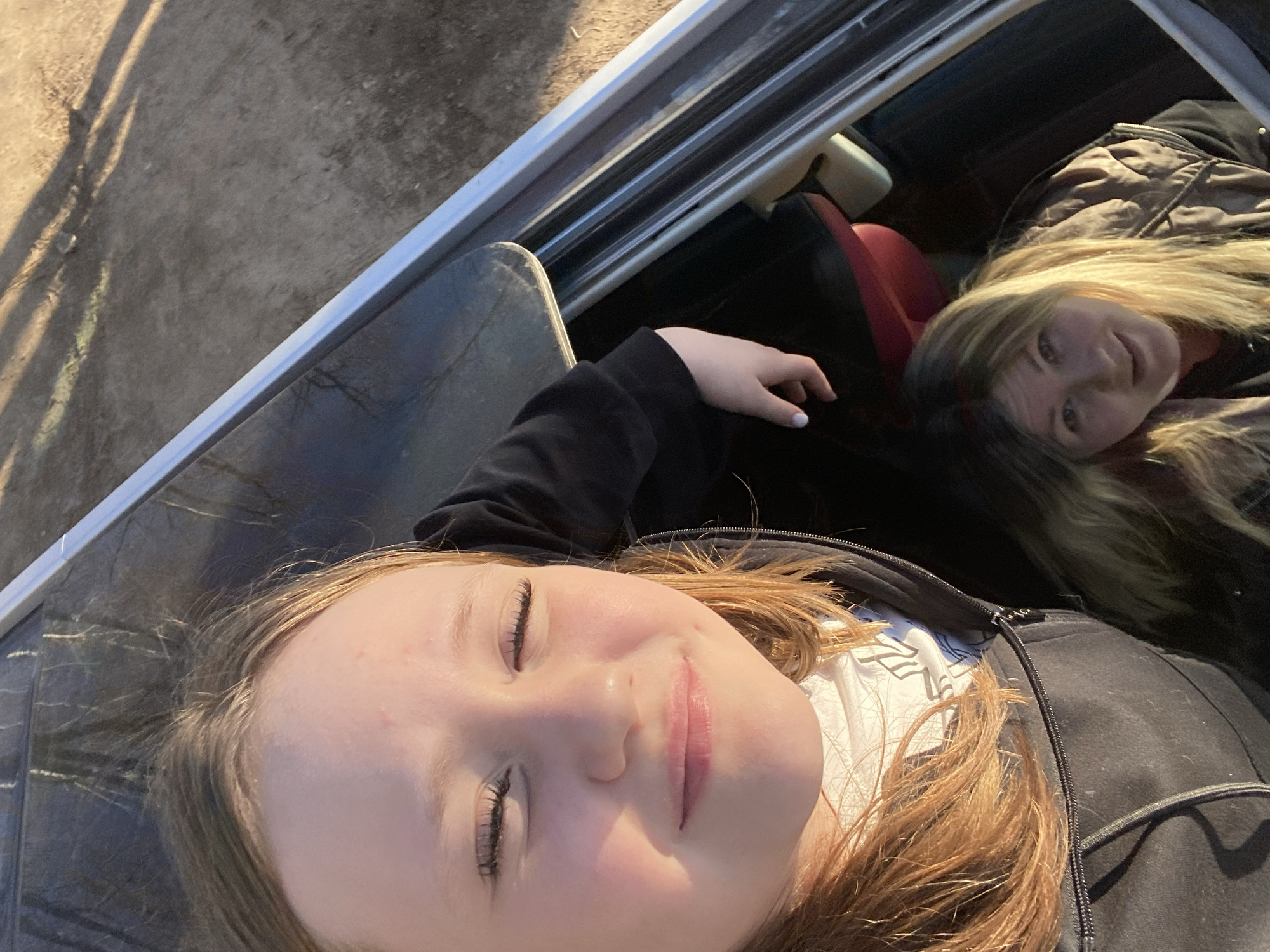 This media file captures two females reclining inside what appears to be a vehicle, likely a car or truck, during daytime. The image is taken from a low angle, looking upwards towards the subjects and the outside environment.
In the foreground, a young female, possibly a child or teenager, lies with her eyes closed and a serene expression, a slight smile on her lips. She has fair skin and light brown or blonde hair, which is illuminated by bright sunlight. She is wearing a dark, possibly black, zipped jacket over a white shirt with a faint pattern. Her left arm is visible, resting by her side, with light-colored nail polish.
In the background, an adult female is visible, positioned further into the vehicle. She is looking directly at the camera with a calm, gentle expression and a slight smile. She has blonde hair and is wearing a patterned jacket, possibly brown or black.
The immediate setting is the interior of a vehicle, with parts of a car seat (red and black upholstery) and interior panels visible. A metallic rail or frame element runs along the upper part of the image, separating the vehicle interior from the exterior. Outside the vehicle, on the left side of the frame, sun-drenched ground is visible, appearing to be dirt or gravel, with distinct shadows suggesting strong sunlight.
The activity appears to be a moment of rest or relaxation within the vehicle. The bright sunlight indicates it is daytime, possibly morning or late afternoon, given the shadows. The atmosphere is peaceful and relaxed. There is no visible text in the image. While the provided location context is Chaikovsky, Russia, there are no specific visual cues within the image to confirm this geographical location.