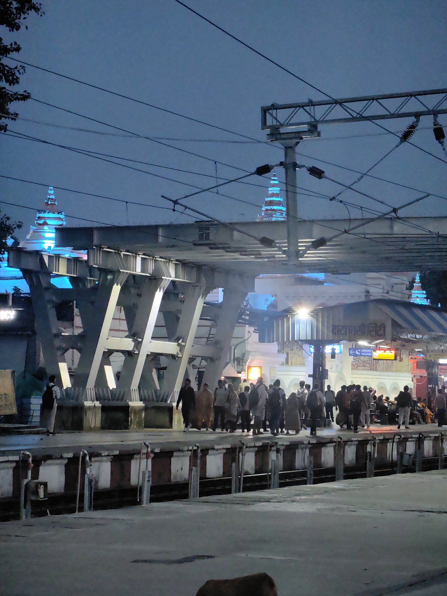 The image depicts a nighttime scene at a train station in Varanasi, India. The sky is a deep blue, indicating dusk or early night. Electrical wires crisscross the frame, supported by metal poles and overhead structures, suggestive of an active railway environment. In the background, illuminated spires, likely part of a temple or monument, glow with bright blue light.
A platform is visible in the foreground, lined with people waiting or walking. They are dressed in various casual attire, some carrying bags. The platform itself has a distinct border with white and dark stripes. The architecture of the station includes a prominent elevated walkway or bridge supported by large, angular metal beams. Signage in Hindi can be observed on the station buildings. The ground in the foreground appears to be a concrete or paved surface, with some debris. The overall atmosphere is that of a busy, public transit hub at night.