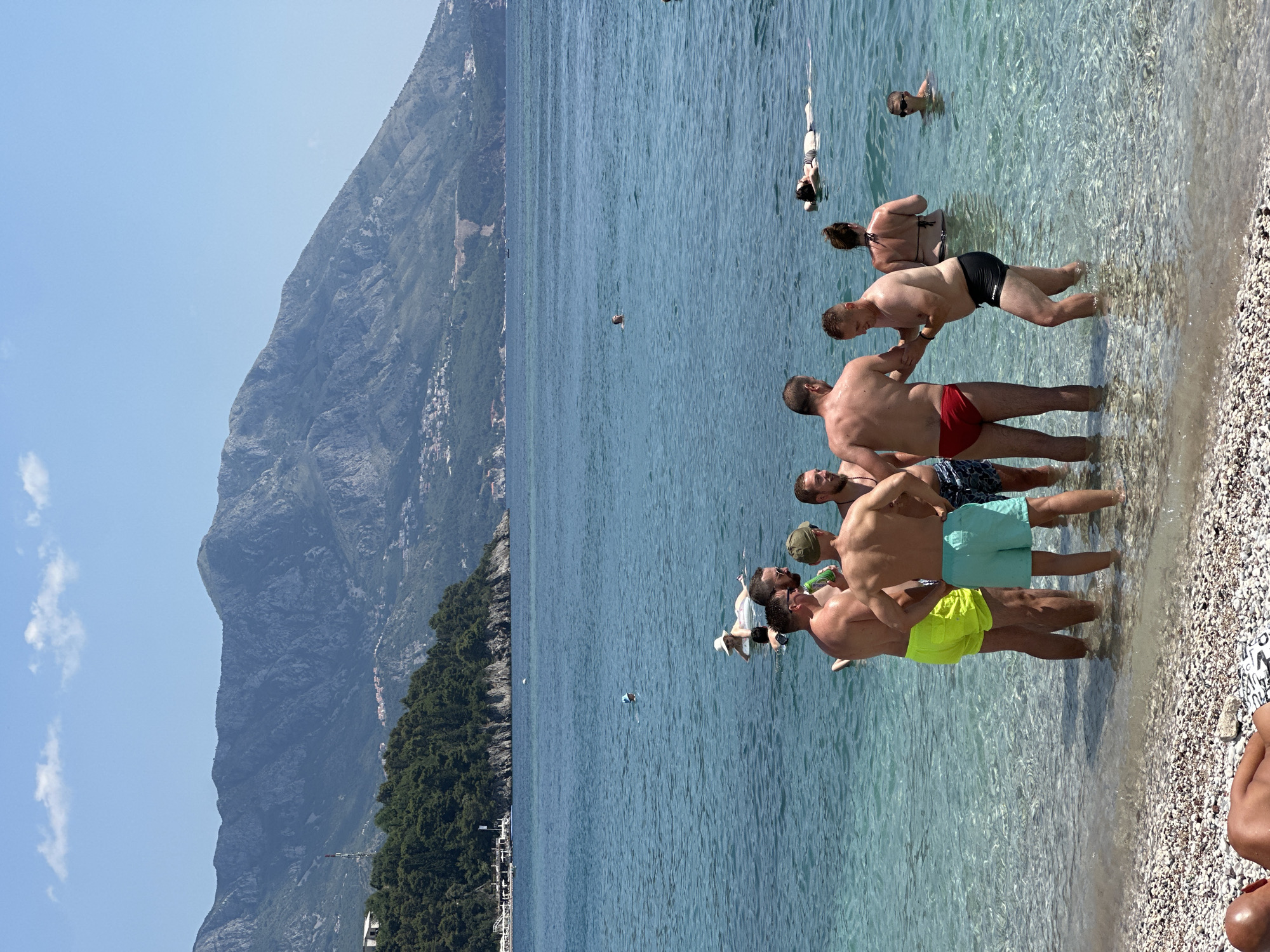The image depicts a sunny day at a beach in Budva, Montenegro. In the foreground, several men are standing in the clear, shallow water, some appearing to be in conversation or interacting. They are wearing swim trunks. Further out in the turquoise water, other individuals are visible, including a woman floating on an inflatable, a person swimming, and another person relaxing on the water. In the background, lush green hills rise steeply, dotted with buildings, and a large, rugged mountain range forms the horizon under a bright blue sky with scattered white clouds. The water is calm and transparent, revealing the rocky seabed near the shore.