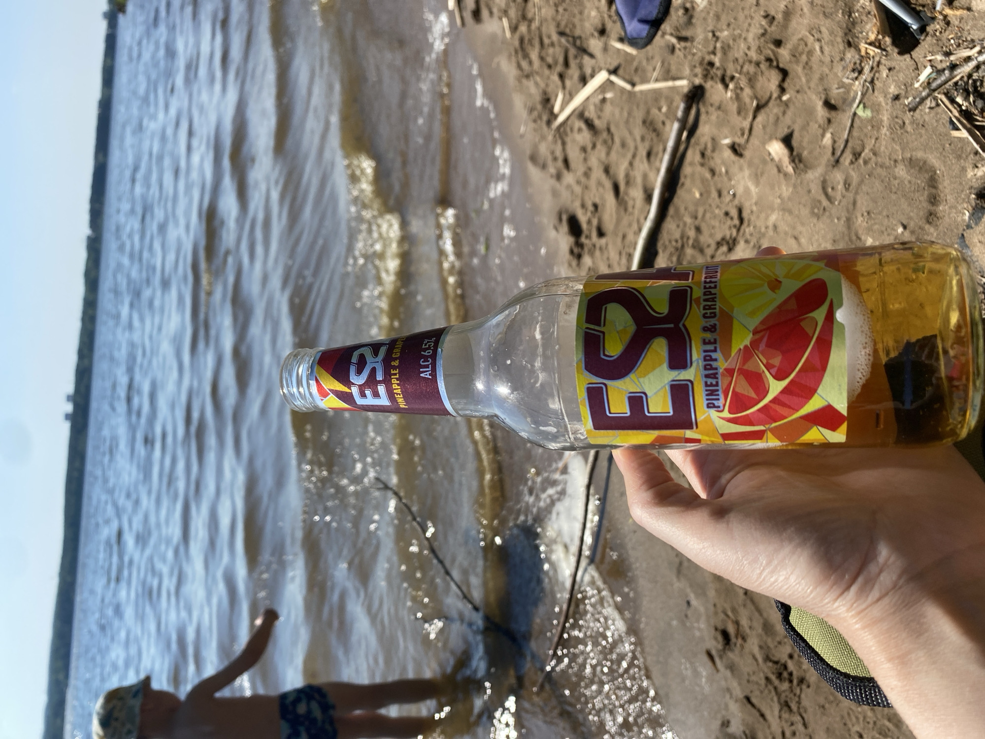 Here is a detailed description of the image:
**Scene:** The image depicts a person holding a bottle of "ES Pineapple & Grapefruit" alcoholic beverage on a beach. A body of water is visible in the background.
**People:** The image shows the arm and hand of an individual holding the bottle. In the background, there is a child.
**Objects:** The primary object is the glass bottle of ES Pineapple & Grapefruit drink. Other objects include a green shoulder strap, sticks, sand, and water.
**Environment:** The location is a beach, as indicated by the sand, water, and possibly a shoreline with trees visible in the distance.
**Location Context:** The bottle of ES Pineapple & Grapefruit drink suggests a possible location within Russia, though it could be anywhere the drink is available. The city of Chaykovsky is not independently verified in this image.