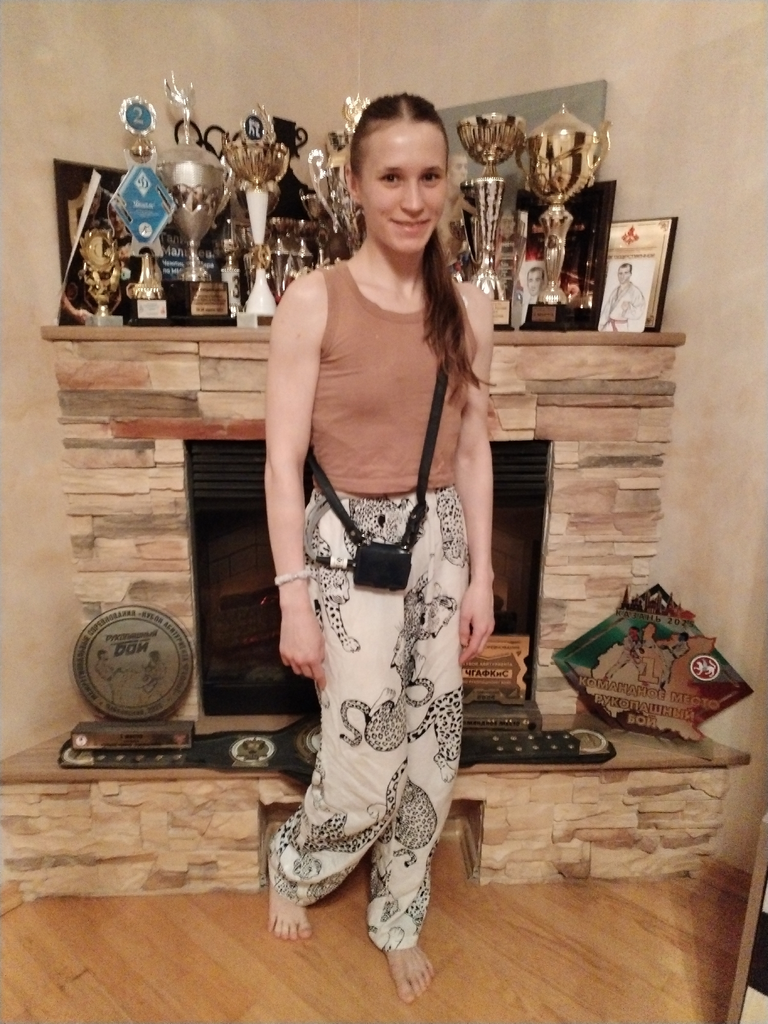 A young woman stands barefoot in front of a stone fireplace mantel, which is heavily adorned with numerous sports trophies, medals, and awards. She is smiling directly at the camera, wearing a plain tan sleeveless top and loose-fitting, light-colored pants with a dark leopard or animal print design. A small black pouch or bag hangs across her body by a strap.
The fireplace serves as a display for a collection of achievements, primarily golden and silver trophies of various sizes. Among the awards, several pieces of text in Cyrillic are visible. One prominent award mentions "Галь Мальцев Чемпи" (Gal Maltsev, Champi-), suggesting a name and championship. Another circular medal or plaque reads "Рукопашный" (Hand-to-hand combat) and "СОРЕВНОВАНИЙ «КУБЛЯ АБИТУРИЕНТА WARRA" (Competitions "Applicant's Cup WARRA"). On the lower left of the mantel, a large, dark championship belt is visible, and on the right, a multi-faceted award displays "КАЗАНЬ 2025" (Kazan 2025), "ЧГАФКИС" (ChGAFKiS, likely Chaykovsky State Academy of Physical Culture and Sport, given the context of Chaikovsky, Russia), "КОМАНДНОЕ MECTO" (Team place), and "РУКОПАШНЫЙ БОЙ" (Hand-to-hand combat fight). Portraits of men in martial arts uniforms are also visible on some framed awards. The setting appears to be an indoor space, likely a home, in Chaikovsky, Russia, showcasing the woman's achievements in martial arts or combat sports.