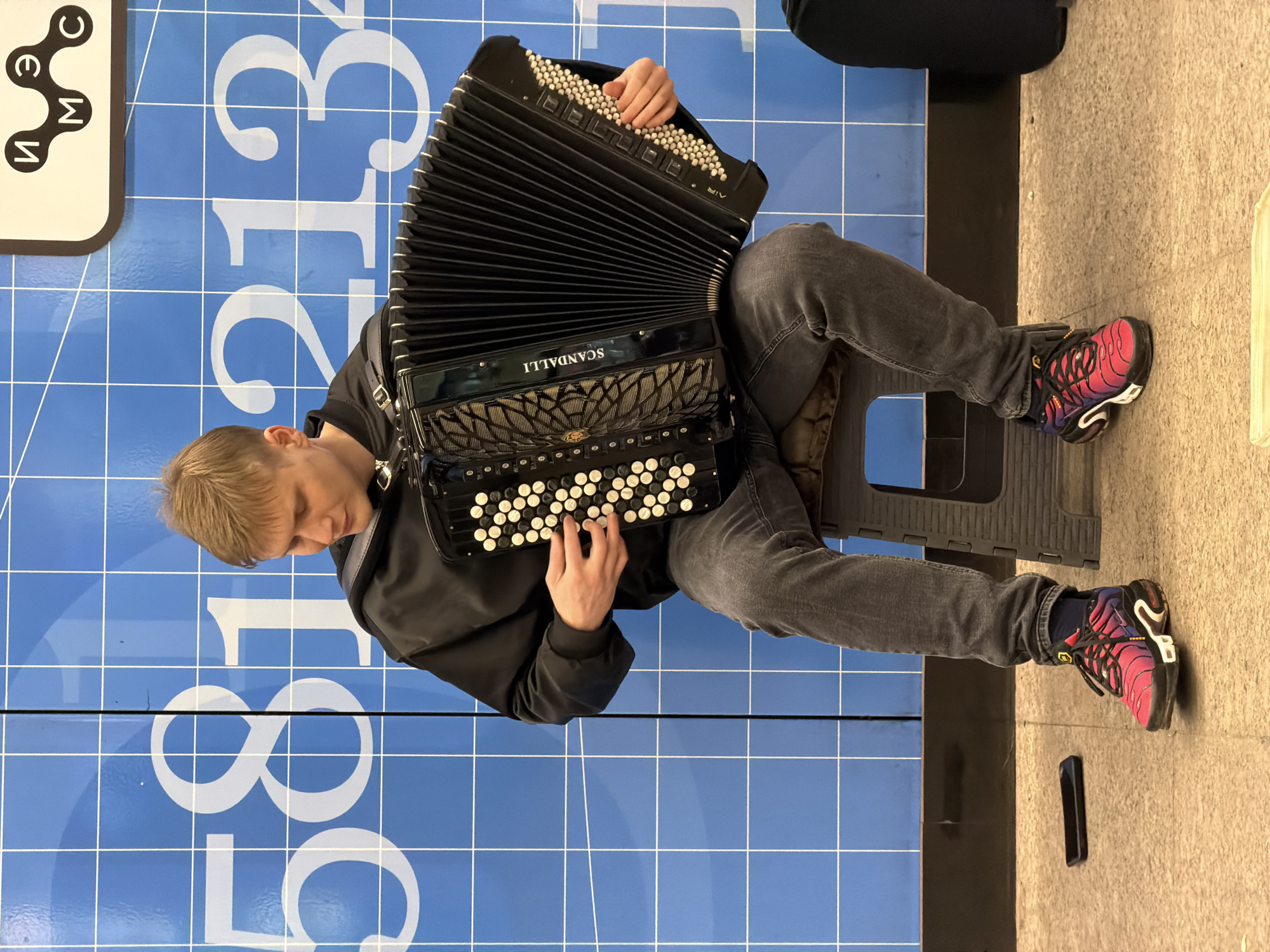A person is seated on a stool and playing a black Scandalli accordion. They are wearing a black jacket, gray jeans, and multi-colored sneakers. The person is facing downwards with their head slightly inclined towards the accordion.
The location is identified as Moscow, Russia by the blue background which includes white grid lines. The Cyrillic characters "И" "М" "Э" "С" are visible in the upper left corner, which are the first letters of the Moscow Institute of Economics, Statistics and Informatics. The numbers 581321 are visible on the blue surface which is also known to be associated with Moscow's subway stations.
