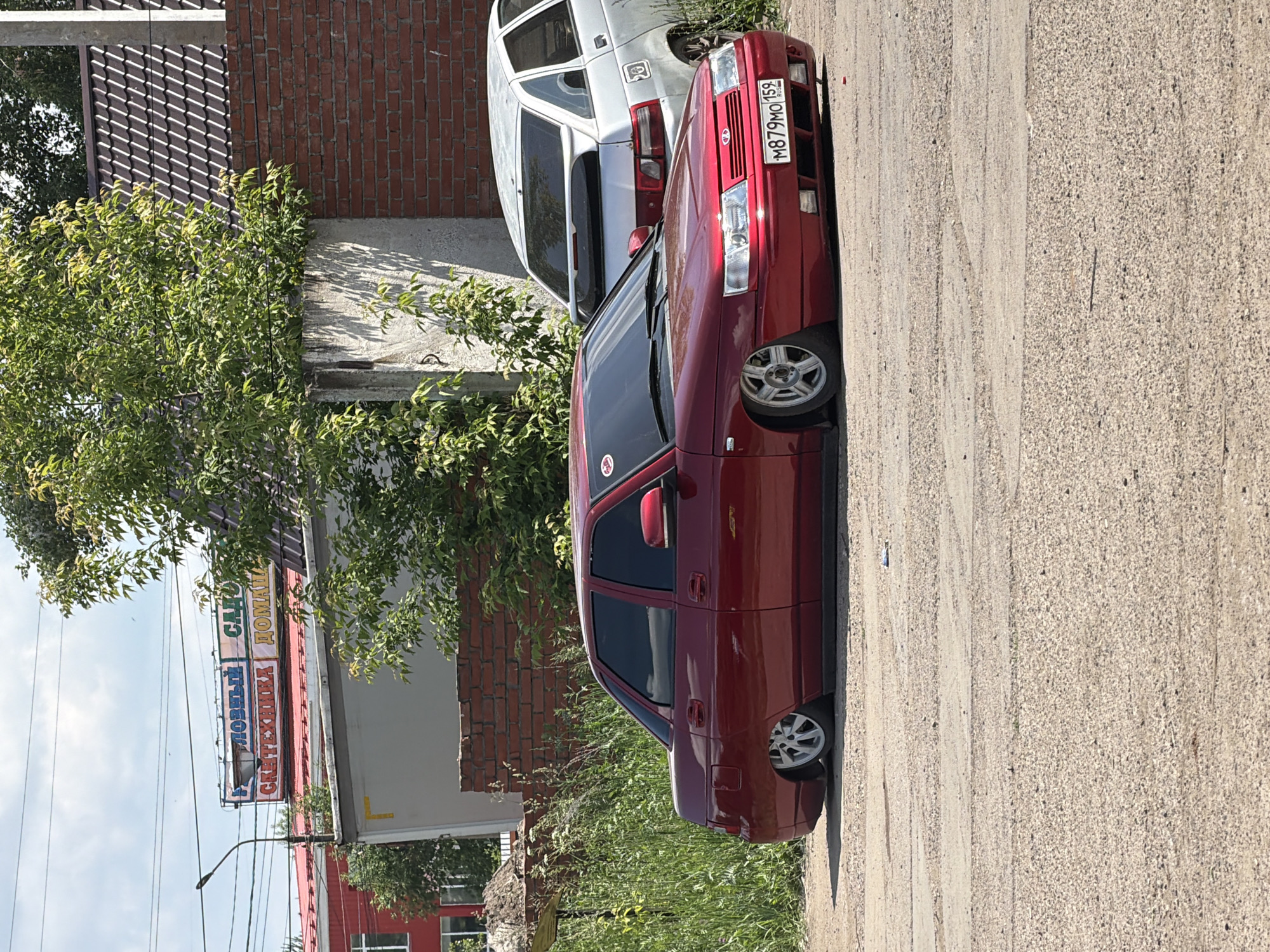 A red sedan is parked on a dirt lot in Chaikovsky, Russia. The car is lowered, with its wheels tucked into the wheel wells. A silver hatchback is parked behind the sedan. In the background, a building with a red roof and a sign that reads "НОВЫЙ САД" (New Garden) is visible. The sky is blue with some clouds.