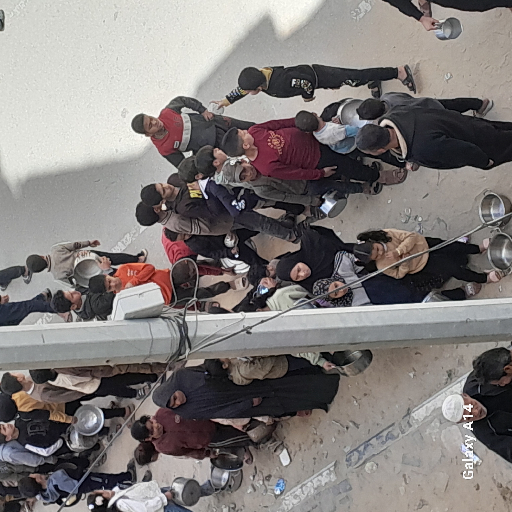 An aerial view shows a long line of people of varying ages, most appearing to be children and young adults, lining up along a street in Unknown, Unknown. Most are holding metal bowls or pots. A few have already been filled. The people are standing or kneeling on a concrete-like surface. Some individuals are wearing head coverings. A utility pole with wiring runs diagonally across the middle ground.