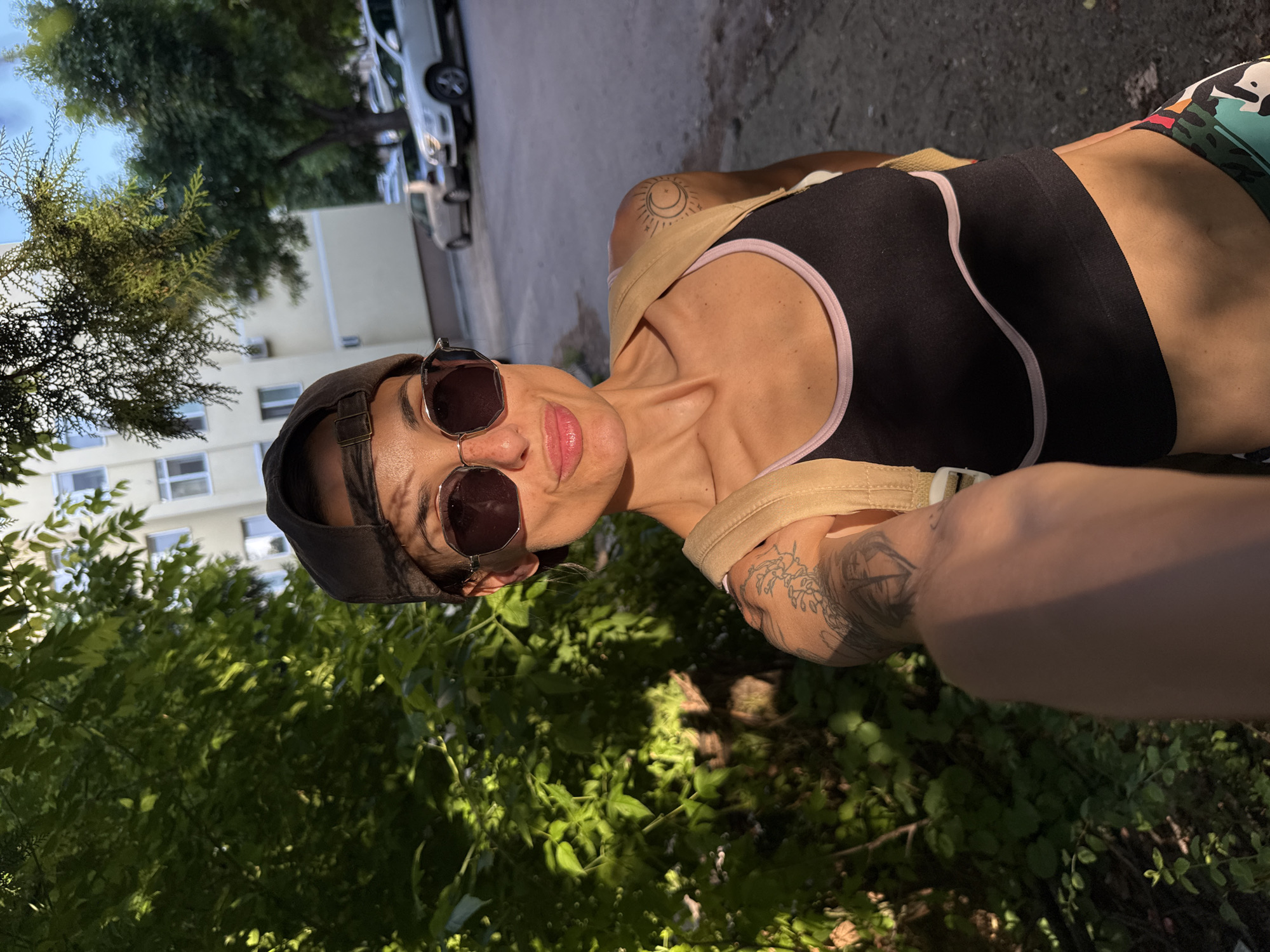 A woman is taking a selfie in what appears to be an outdoor location near Pleven, Bulgaria. She is wearing a black sports bra with a light pink trim, sunglasses, and a baseball cap. She is wearing a brown strap across her chest. Her arm and shoulder are visible, and she has a tattoo on her upper arm. Background vegetation and a building are visible with parked cars and trees in the distance. The lighting suggests it is daytime.